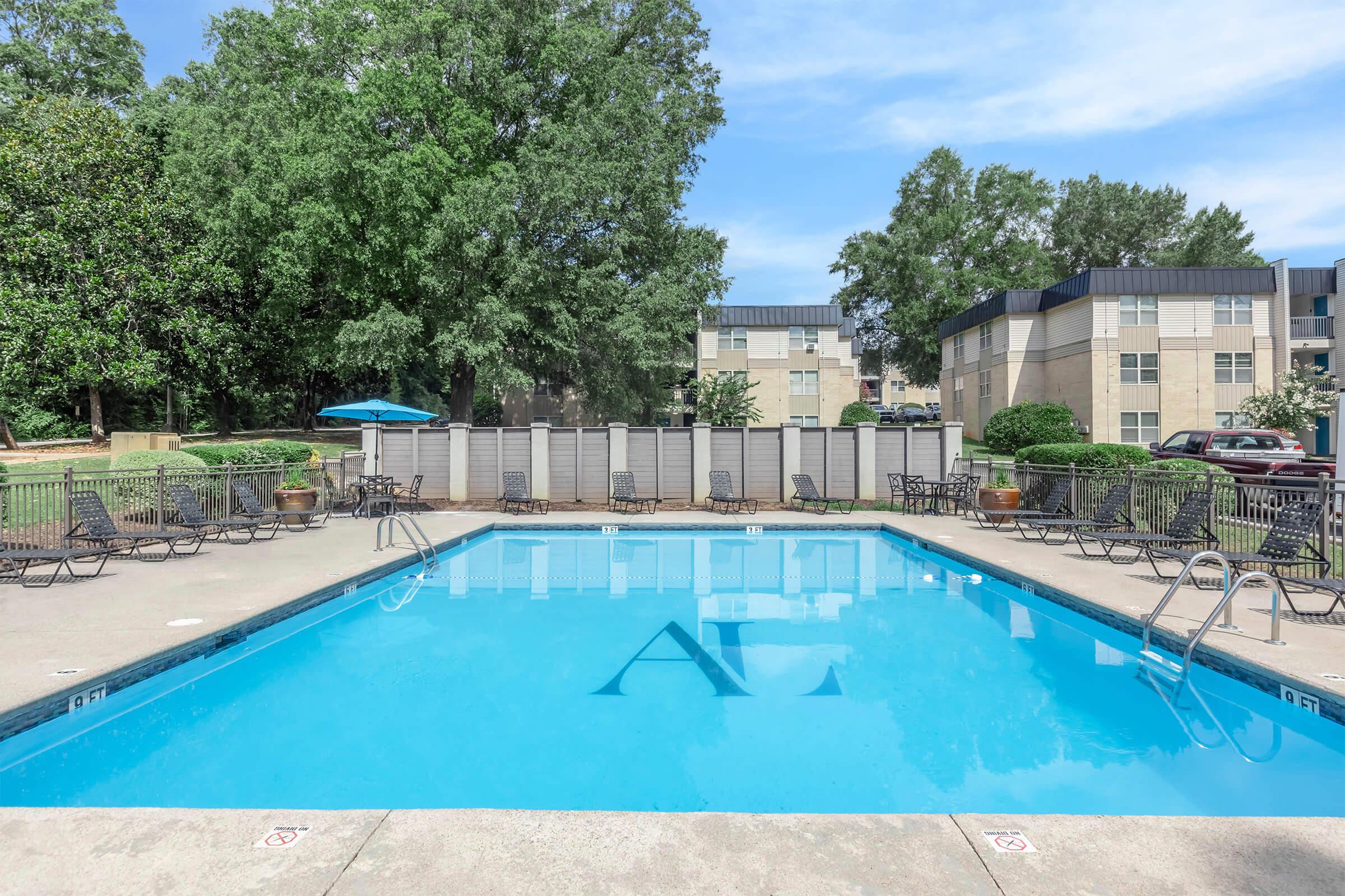 A clear, blue swimming pool surrounded by lounge chairs and small tables, with an umbrella providing shade. In the background, there are trees and apartment buildings with multiple stories under a bright blue sky. The pool area is well-maintained and inviting.