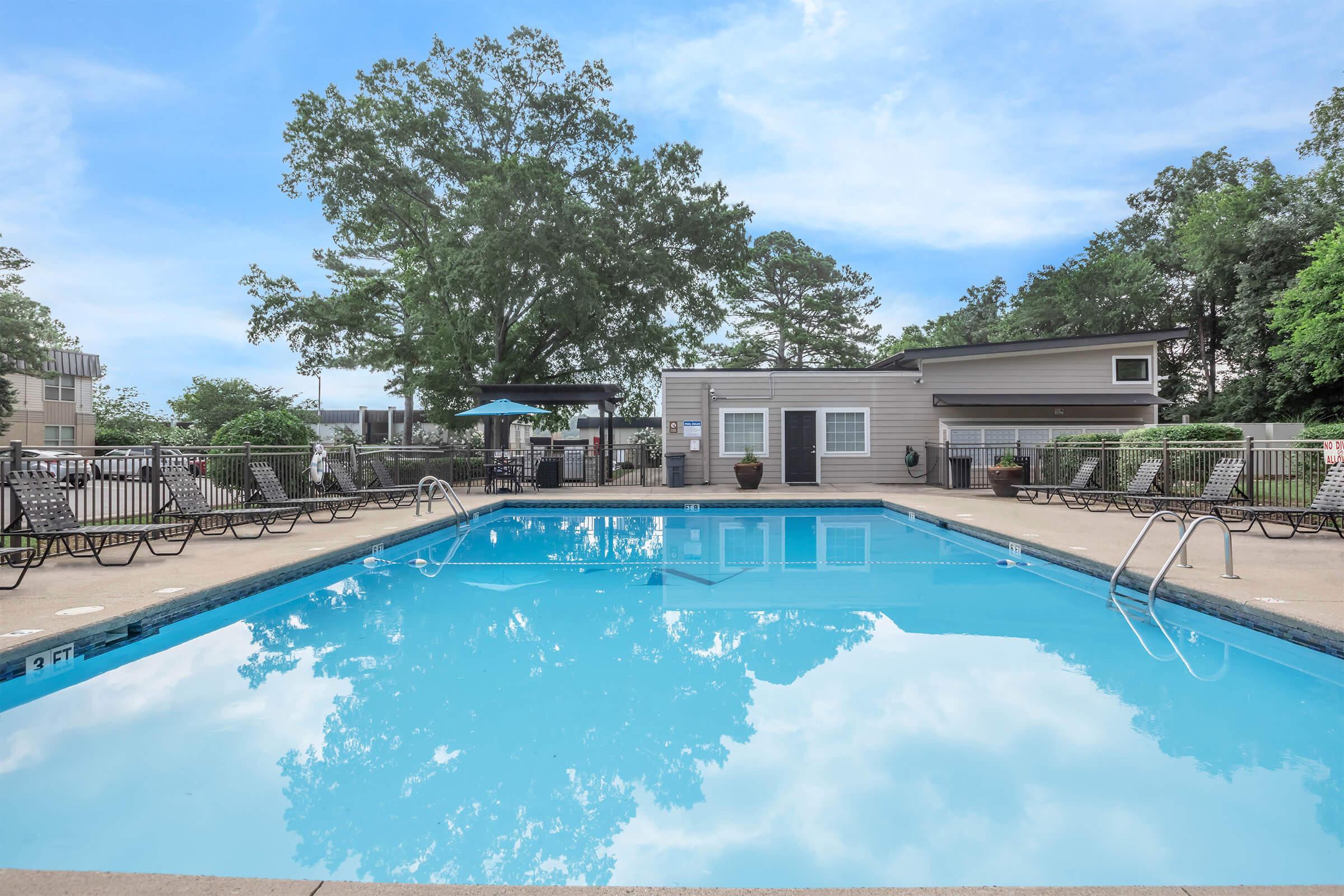 A clear blue swimming pool surrounded by lounge chairs and a fenced area. Green trees and a blue sky in the background. Facilities such as a shaded seating area and a building are visible. The scene conveys a relaxing outdoor atmosphere.