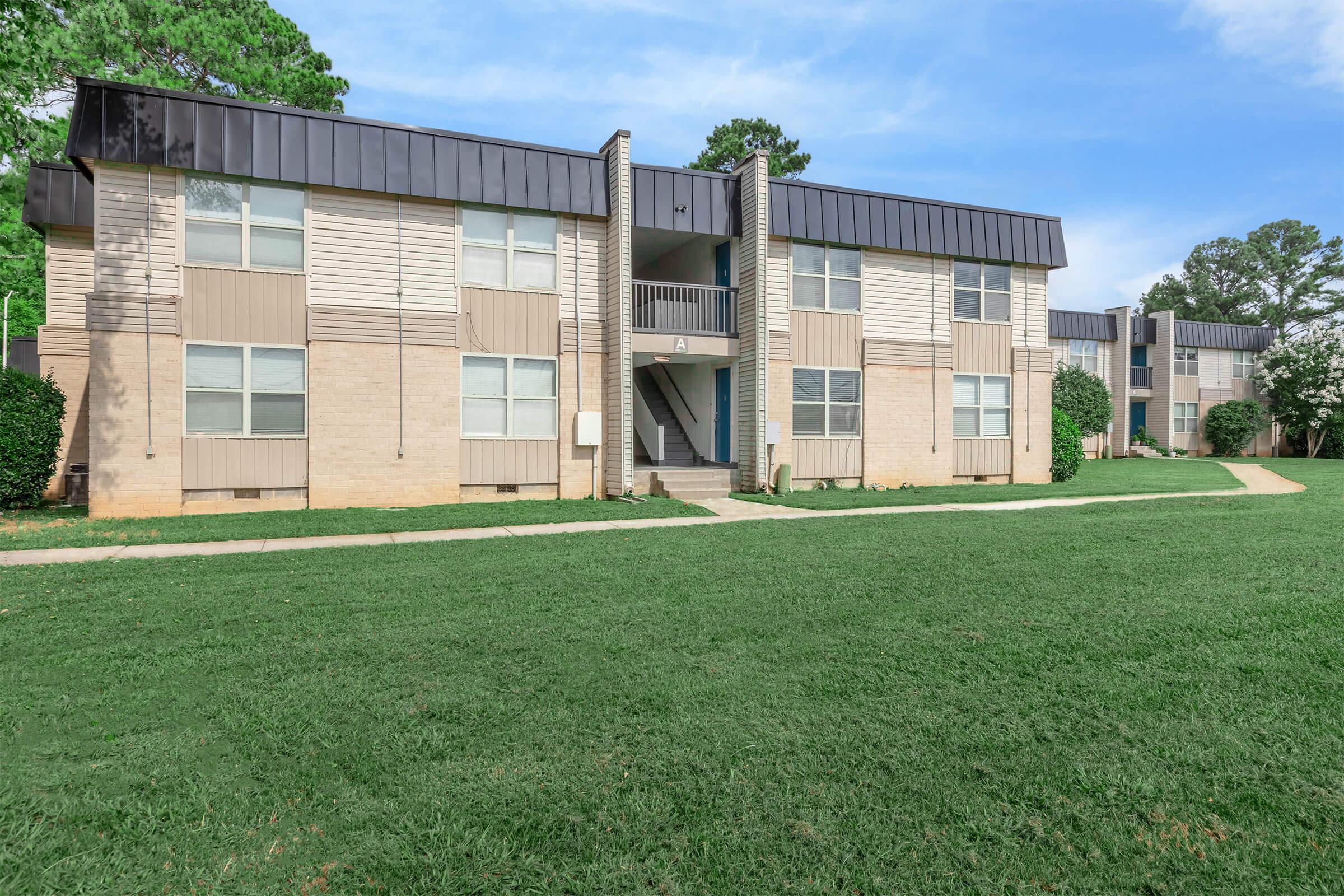Exterior view of a modern two-story apartment building with a light-colored facade, large windows, and a well-maintained lawn. The building features a walkway leading to the entrance and is surrounded by trees and greenery under a clear blue sky.
