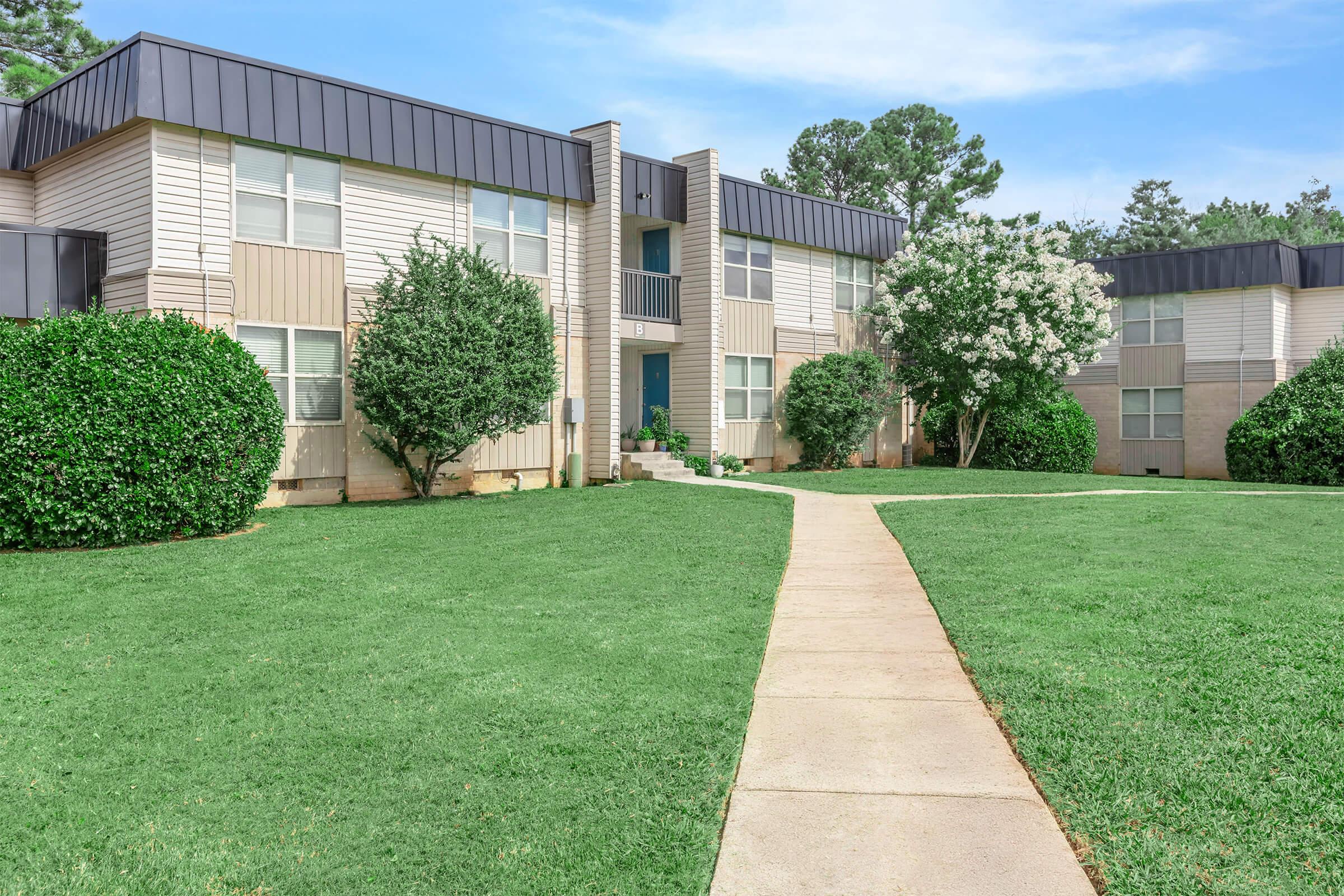 A well-maintained apartment complex featuring two-story buildings with a mix of beige and gray exterior. Lush green lawns surround the building, and neatly trimmed shrubs add to the landscape. A paved path leads to the entrance, framed by trees and blooming flowers in the vicinity.