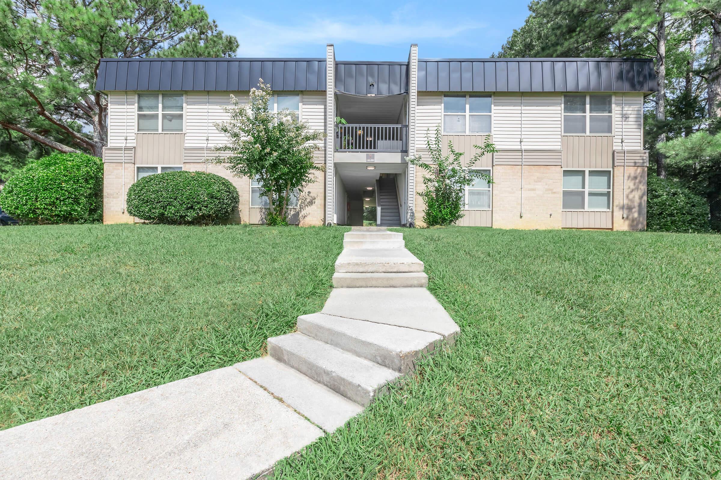 A two-story apartment building with a light beige and gray exterior. There are shrubs on either side and a well-kept lawn in front. A concrete walkway leads to the entrance, which is flanked by small trees. The sky is clear and blue, adding to the bright and inviting atmosphere of the scene.