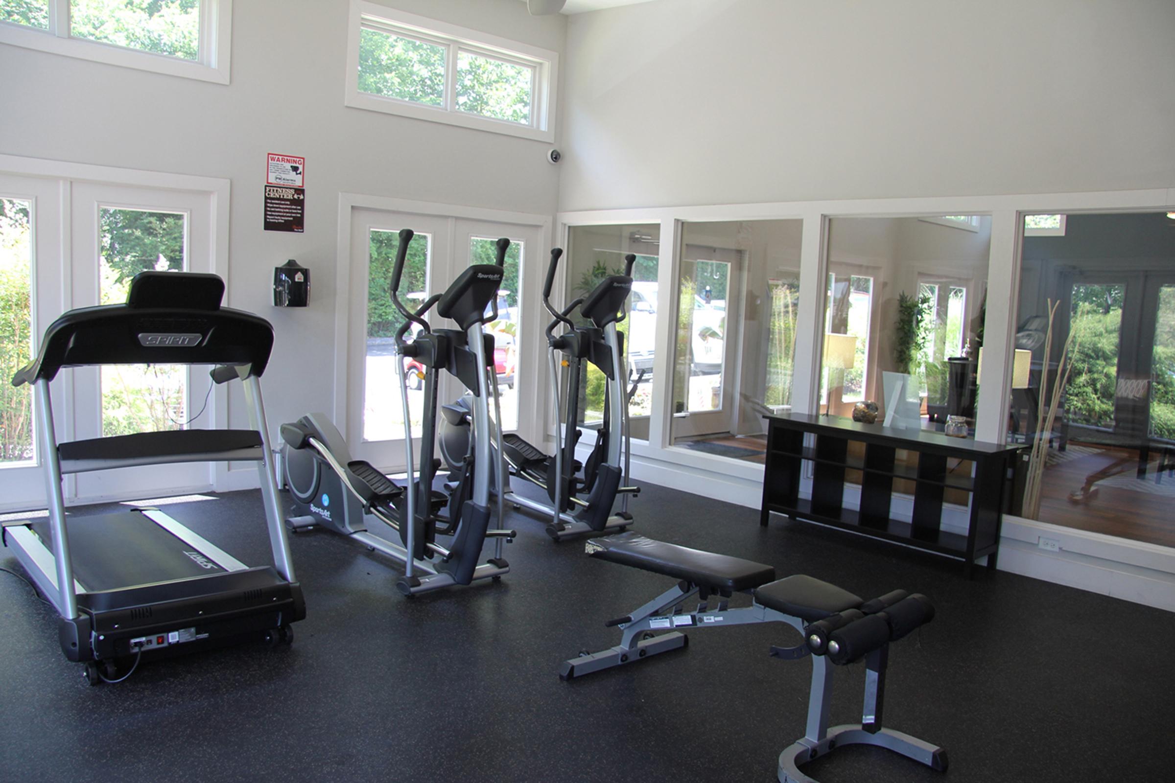A bright and modern gym space featuring a treadmill, an elliptical machine, and a weight bench. The room has large windows allowing natural light to enter, with a mirrored wall reflecting the equipment. A sleek, dark console table is positioned against one wall.