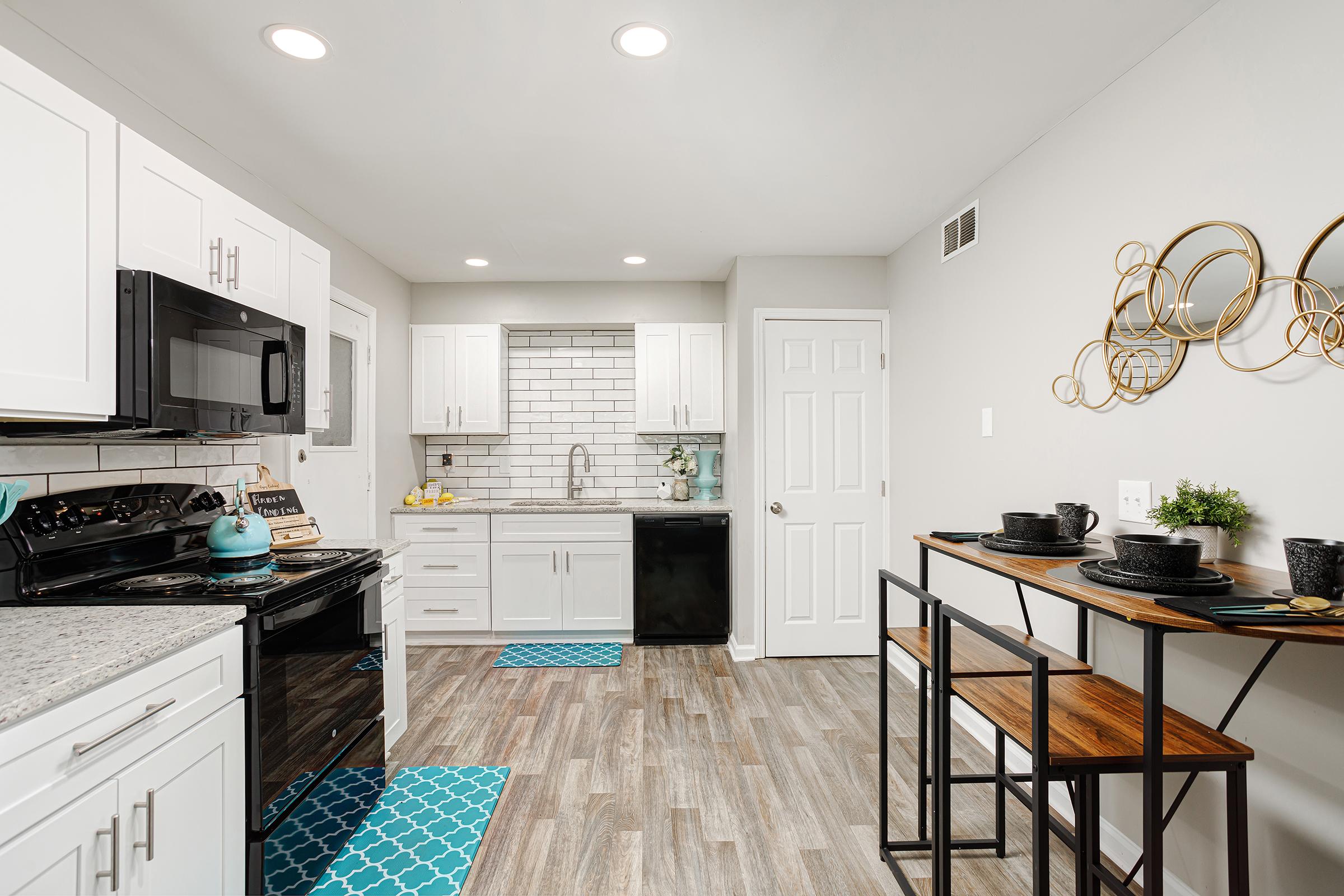 A modern kitchen featuring white cabinetry, stainless steel appliances, and a gray backsplash. The countertops are gray with a granite finish, and a small dining space with two stools is set up. Aqua-colored rugs add a pop of color to the floor. The walls are light gray, creating a bright, airy atmosphere.
