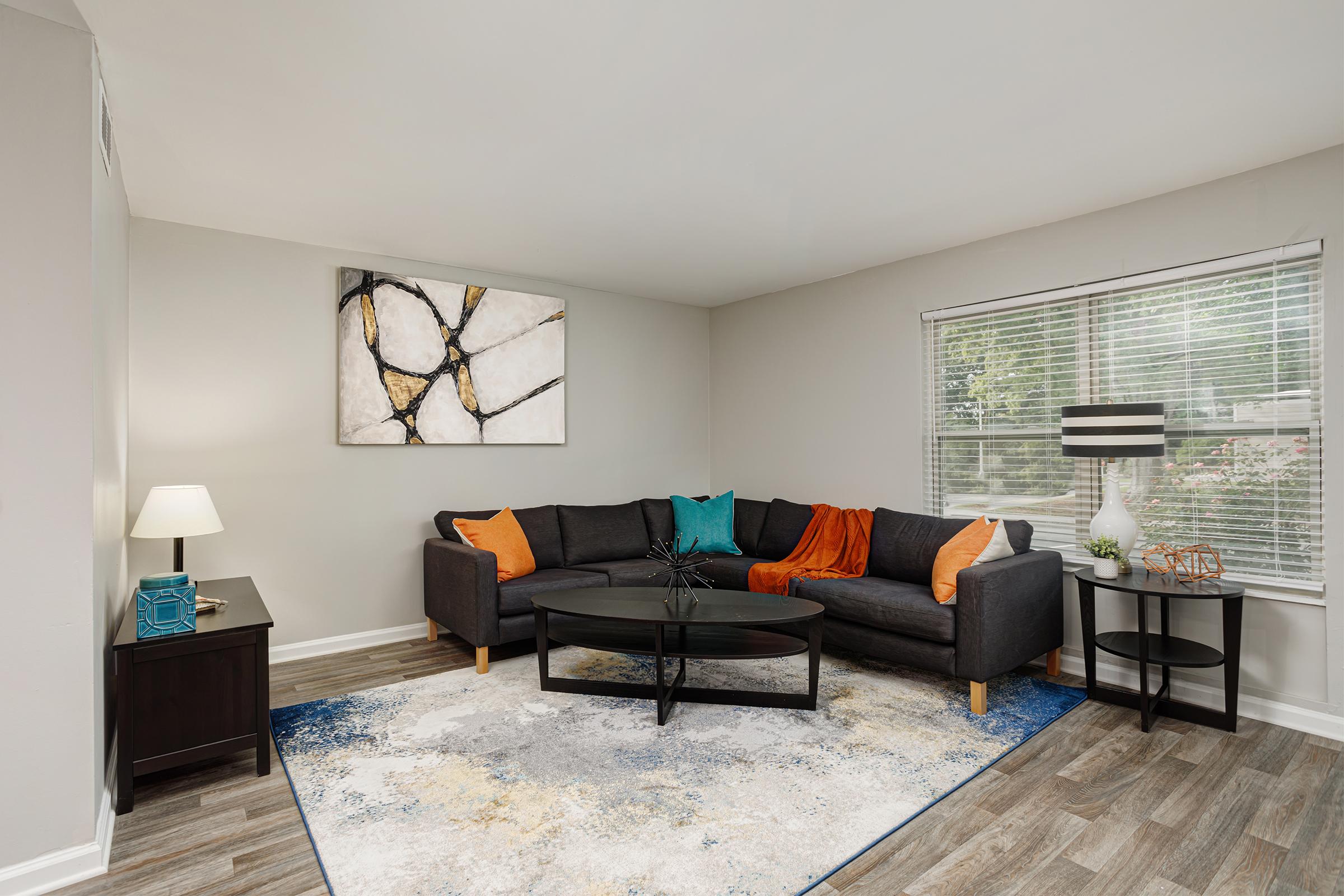 A cozy living room featuring a dark grey sectional sofa adorned with colorful pillows and a vibrant orange throw. There is a round black coffee table in front of the sofa, an abstract wall art piece above it, a modern lamp on a side table, and large windows allowing natural light to illuminate the space.