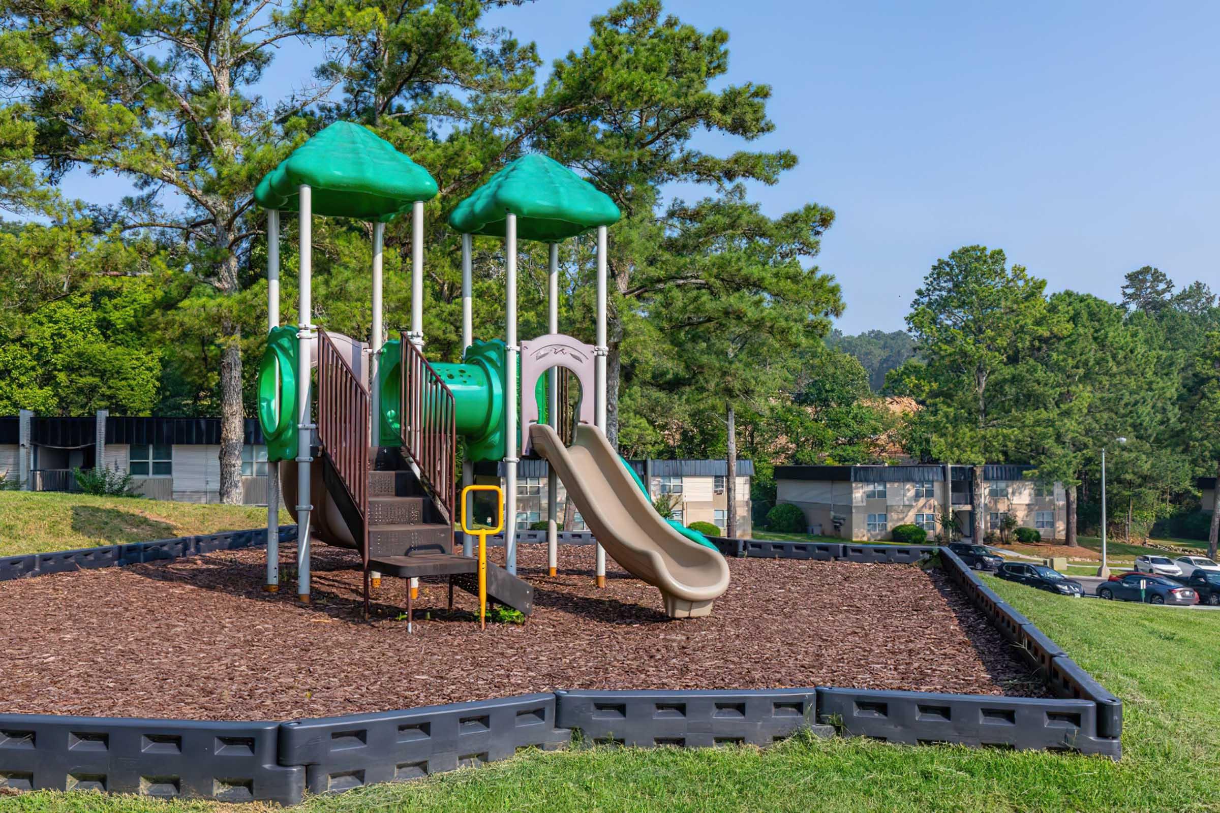A playground setup featuring two play structures with green roofs, a slide, and steps, surrounded by mulch and grassy areas. In the background, there are trees and a parking lot with cars. The scene is bright and sunny, indicating a pleasant outdoor environment for children to play.