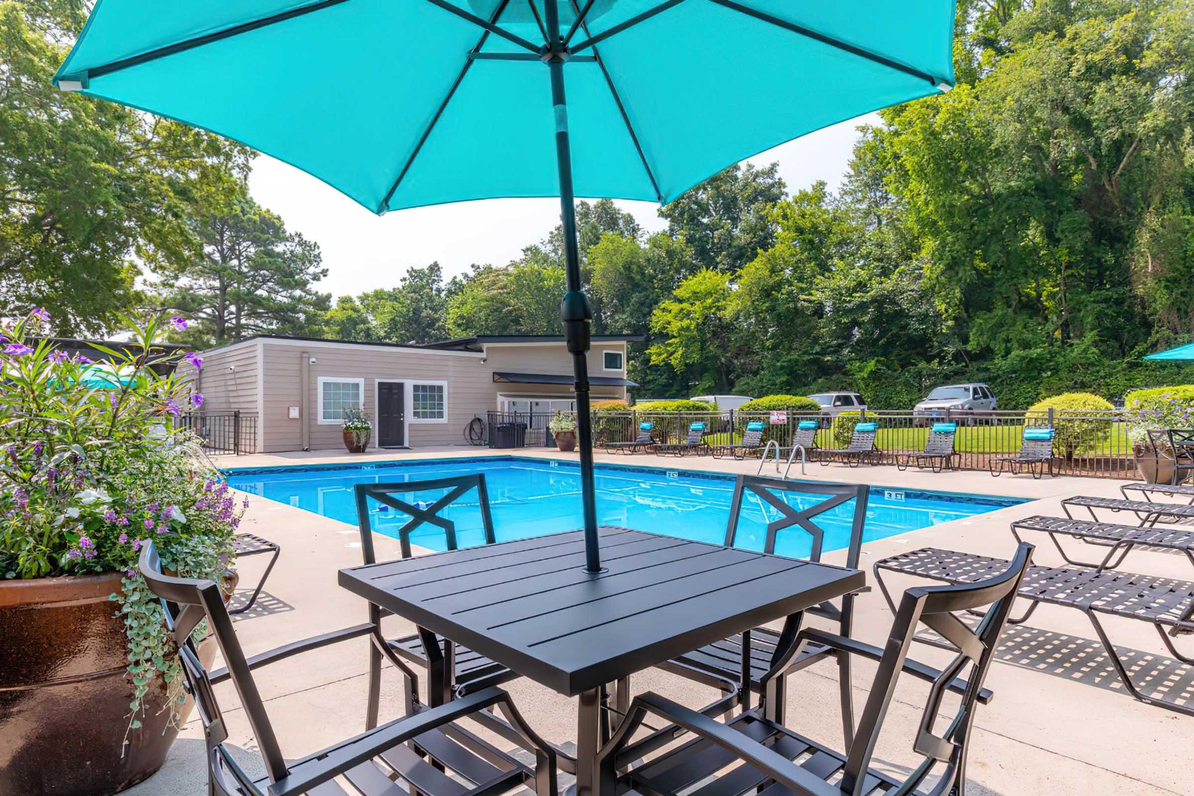 A swimming pool surrounded by lounge chairs and tables with a turquoise umbrella. Lush greenery in the background provides a serene atmosphere, while a few parked cars are visible near the pool area. The scene is bright and inviting, ideal for relaxation and leisure.