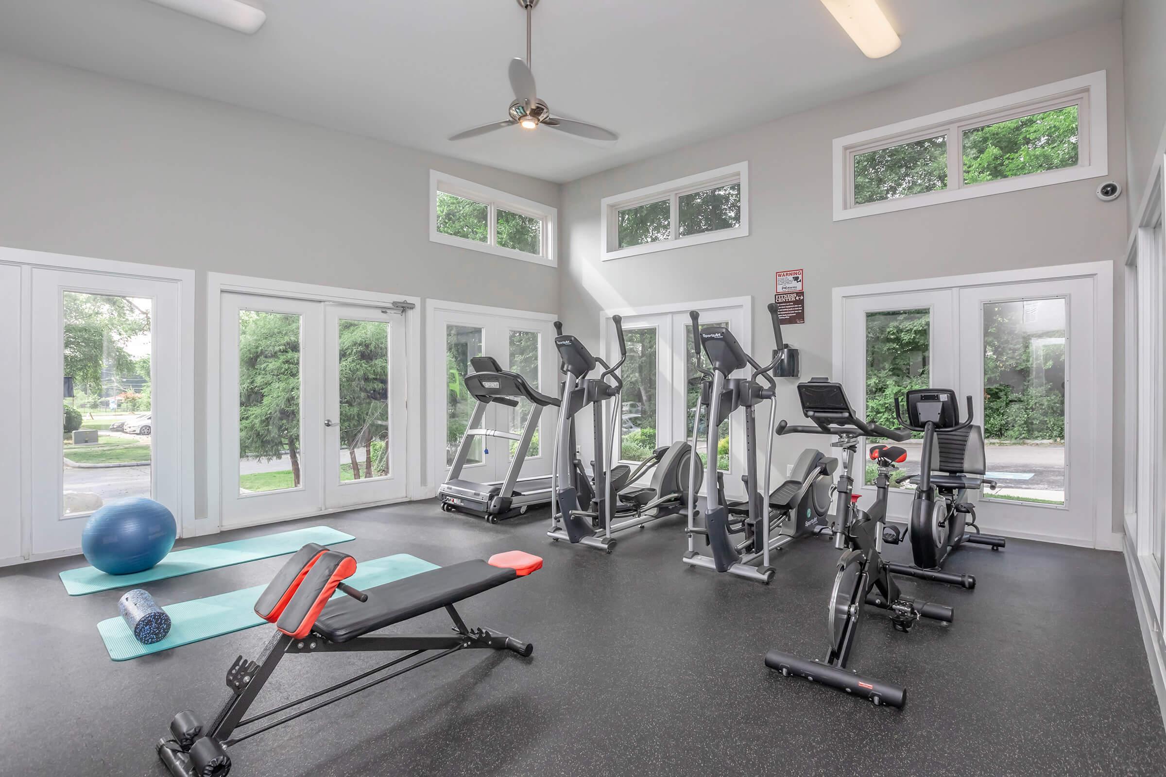 A bright fitness room with large windows, featuring gym equipment such as treadmills, stationary bikes, and a weights bench. Exercise mats and a fitness ball are on the floor, creating an inviting space for workouts.