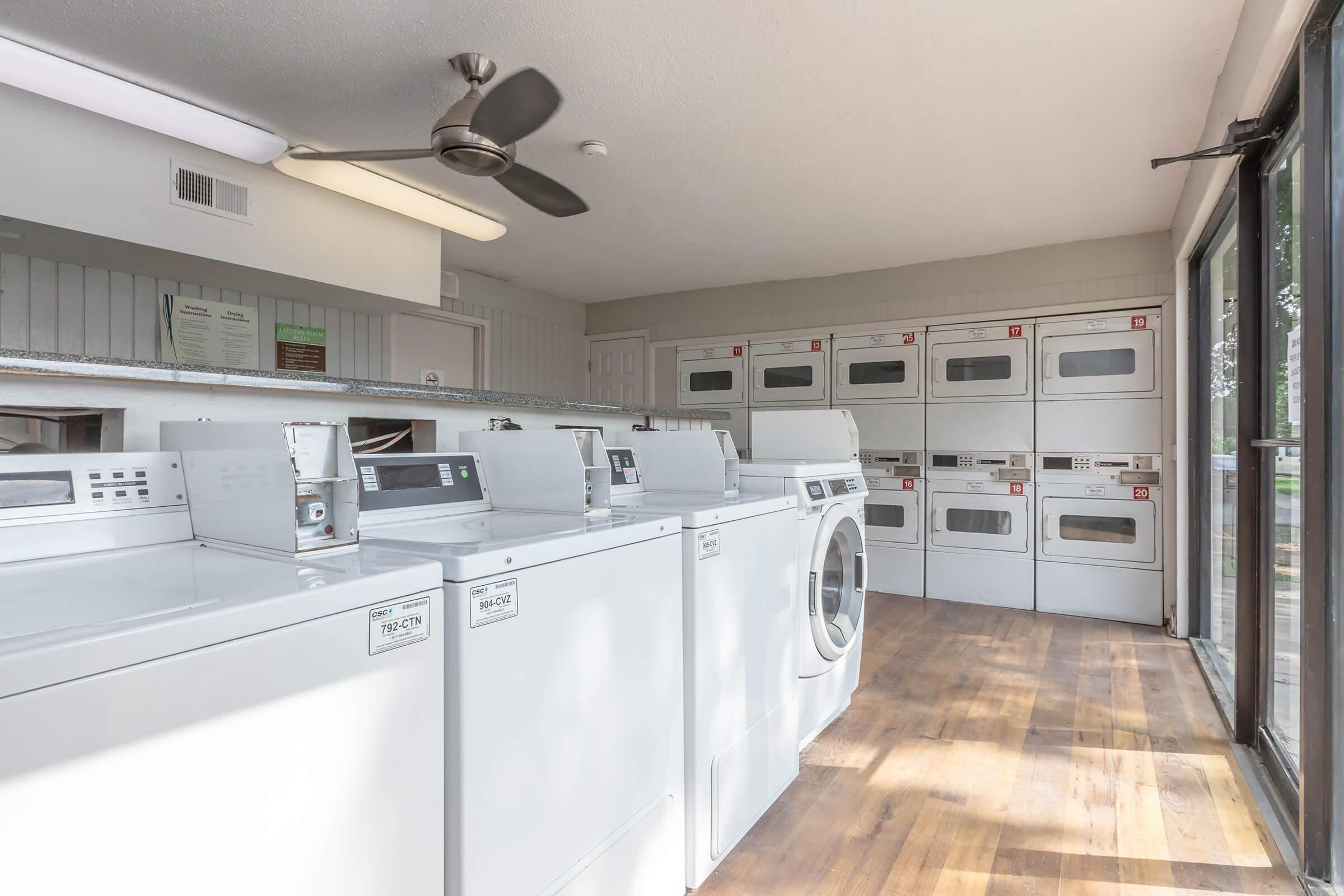 A bright laundry room featuring rows of washing machines and dryers, with a ceiling fan overhead. Large windows allow natural light to fill the space, highlighting the wooden flooring. Wall-mounted cabinets in the background provide storage for laundry supplies.