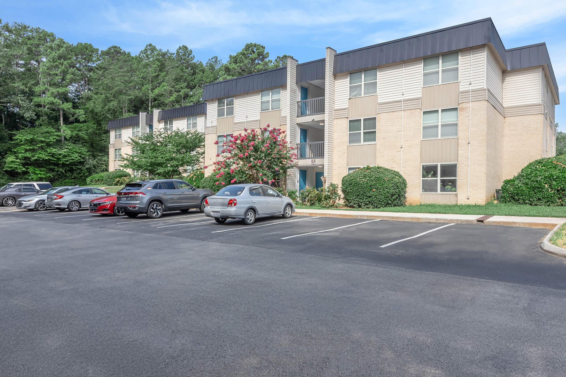 A two-story apartment building with a light brick exterior and a dark roof. The building is surrounded by green trees and shrubs. Several parked cars are visible in the foreground, and the parking lot has multiple empty spaces.