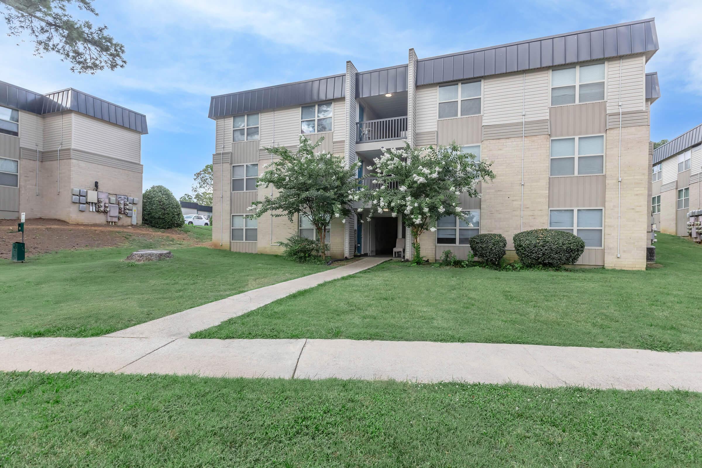 Three-story apartment building with a light-colored facade, featuring balconies and landscaped green spaces. A paved walkway leads to the entrance, flanked by bushes and trees, with additional buildings visible in the background. The sky is partly cloudy, adding a serene atmosphere to the scene.