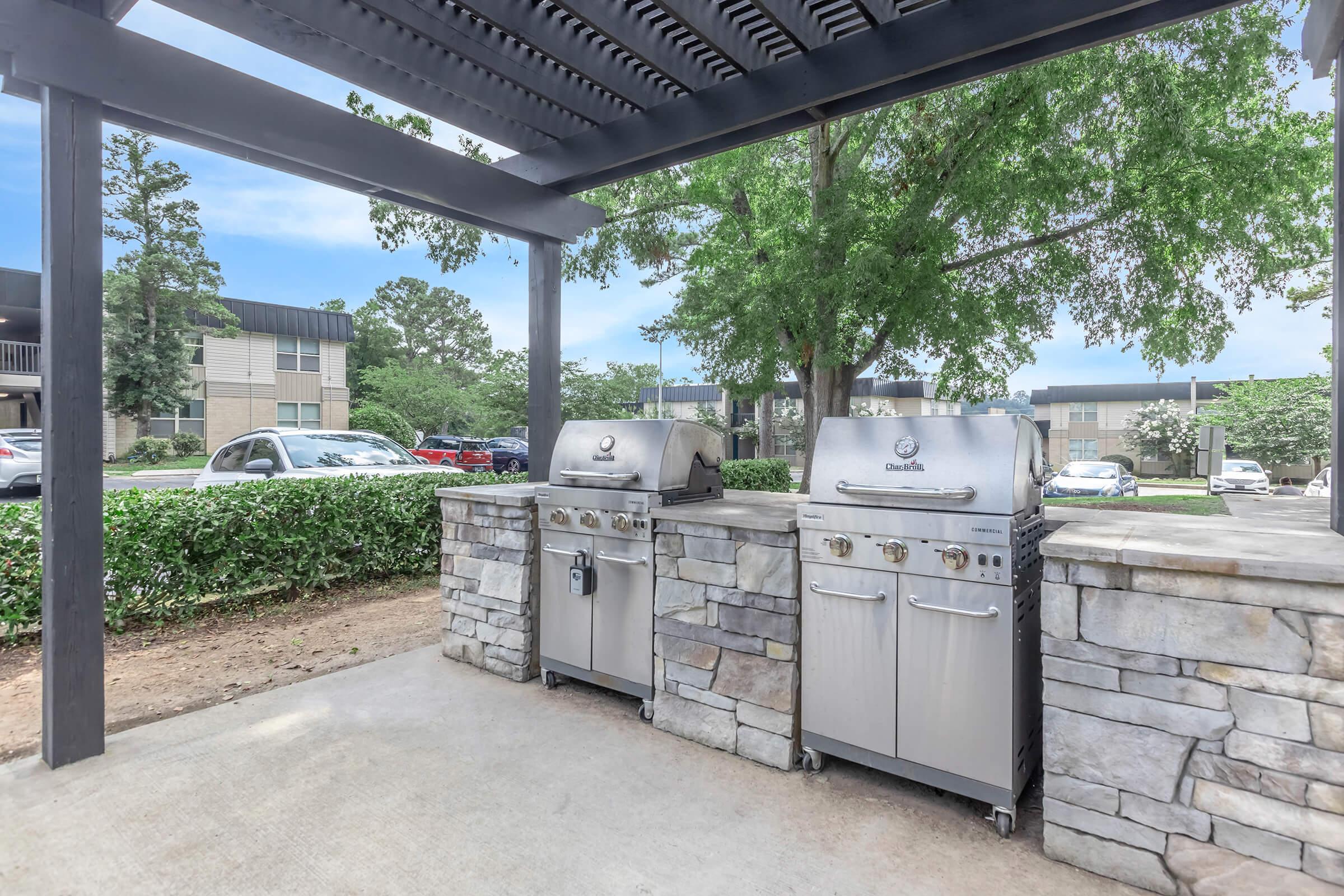 Two stainless steel grills located under a shaded pergola, set against a backdrop of greenery and an apartment complex. A stone countertop supports the grills, creating an outdoor cooking area. The setting is calm and inviting, ideal for barbecues or gatherings.