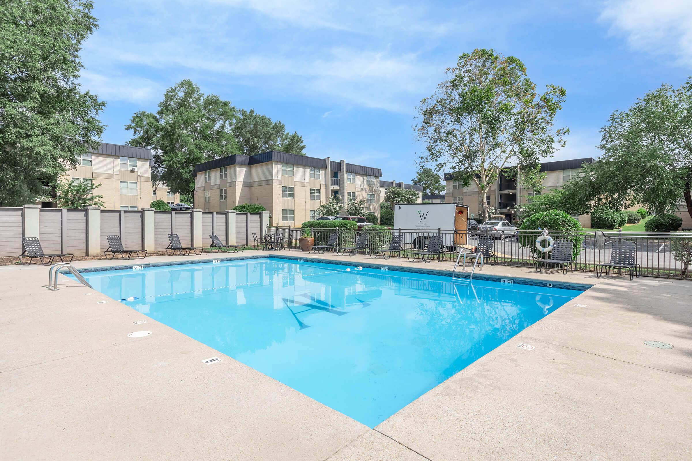 A clear blue swimming pool surrounded by a concrete deck, with several lounge chairs along the sides. In the background, there are multi-story apartment buildings and lush green trees, under a clear blue sky. The area is well-maintained and inviting for residents.