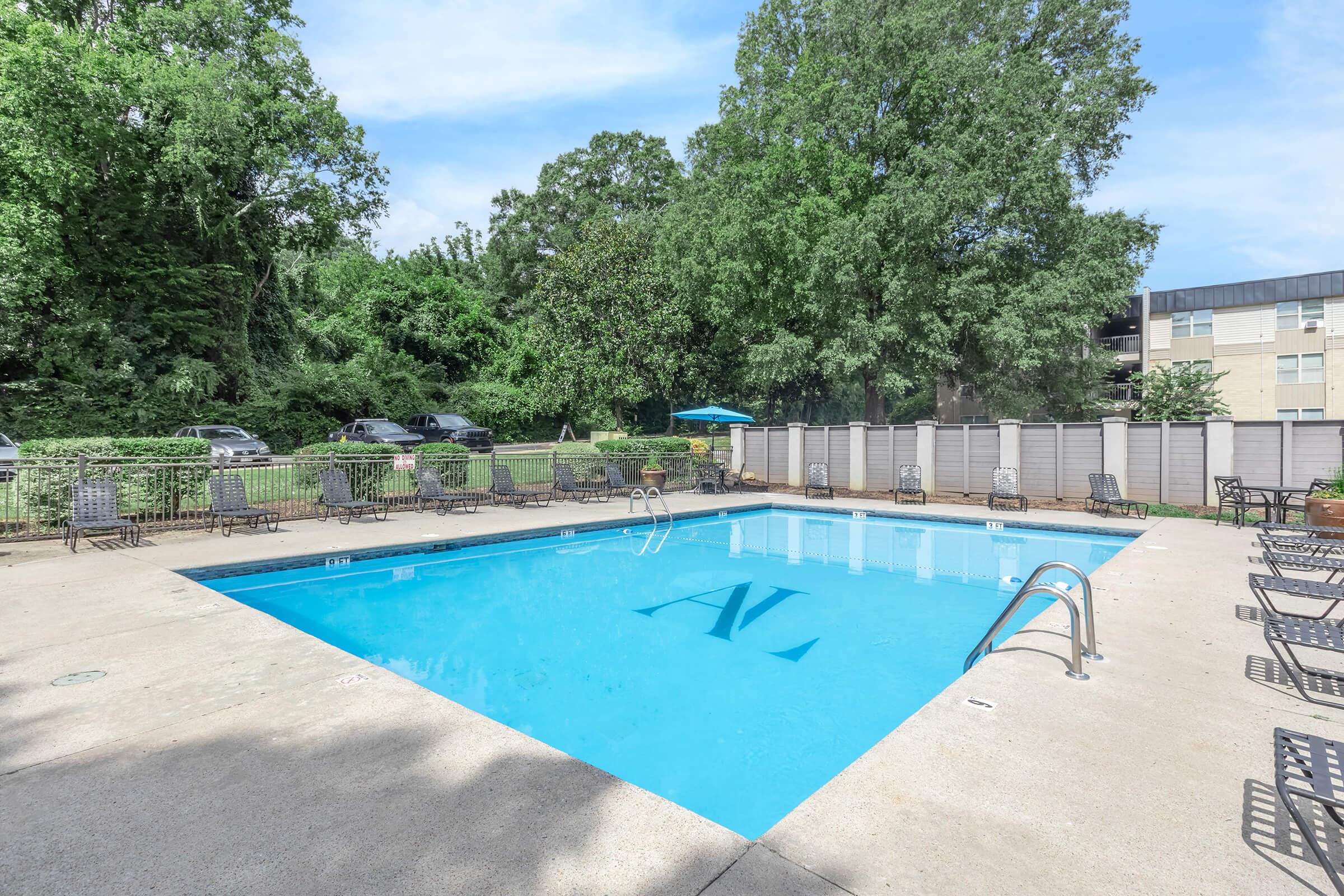A clear blue swimming pool surrounded by a concrete deck. Lounge chairs are arranged around the pool, and there is a shaded area with an umbrella. Lush green trees are visible in the background, providing a tranquil setting.