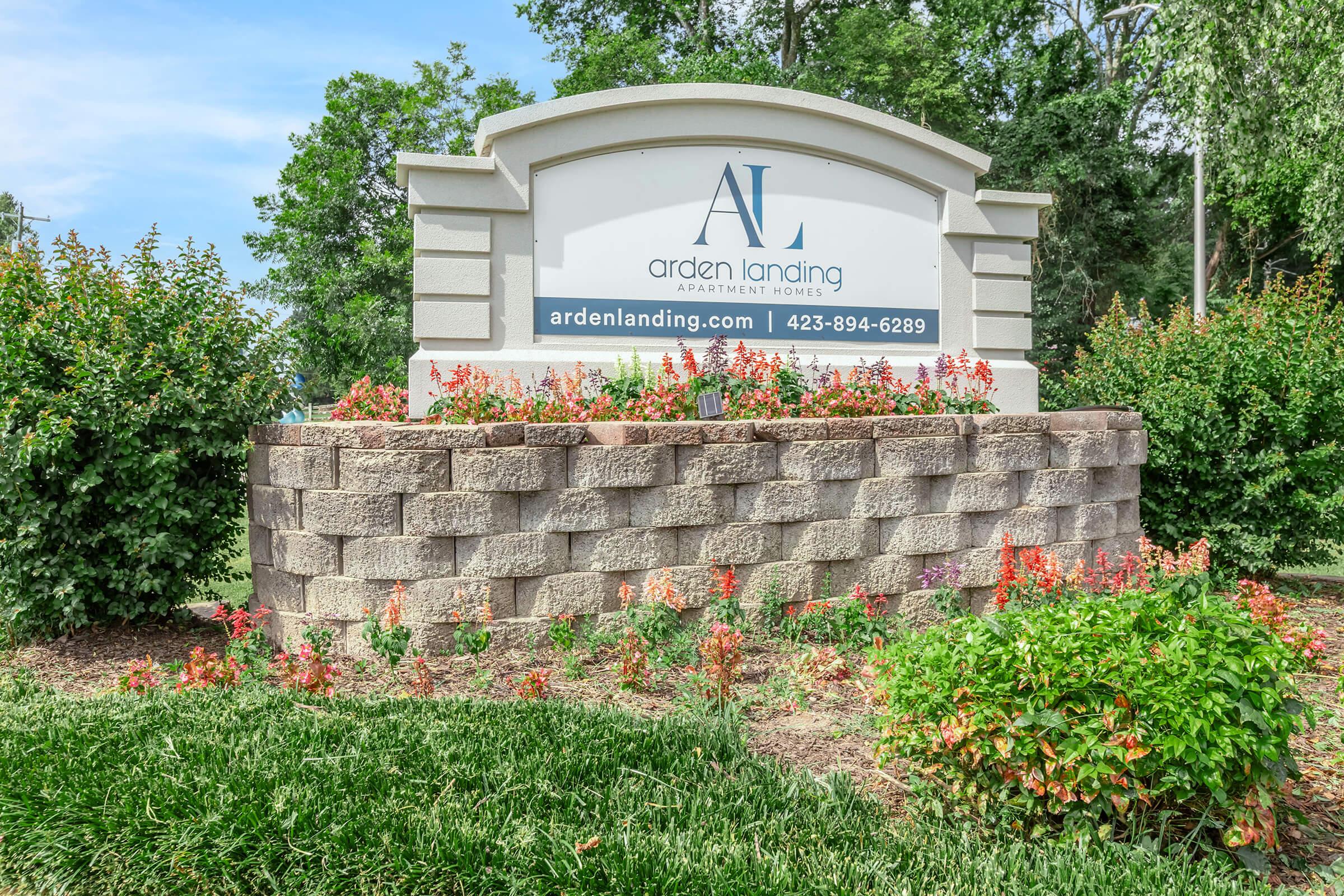 Sign for Arden Landing Apartment Homes featuring the website ardenlanding.com and a phone number, surrounded by landscaped flowers and greenery. The sign is prominently displayed against a background of trees and shrubs.