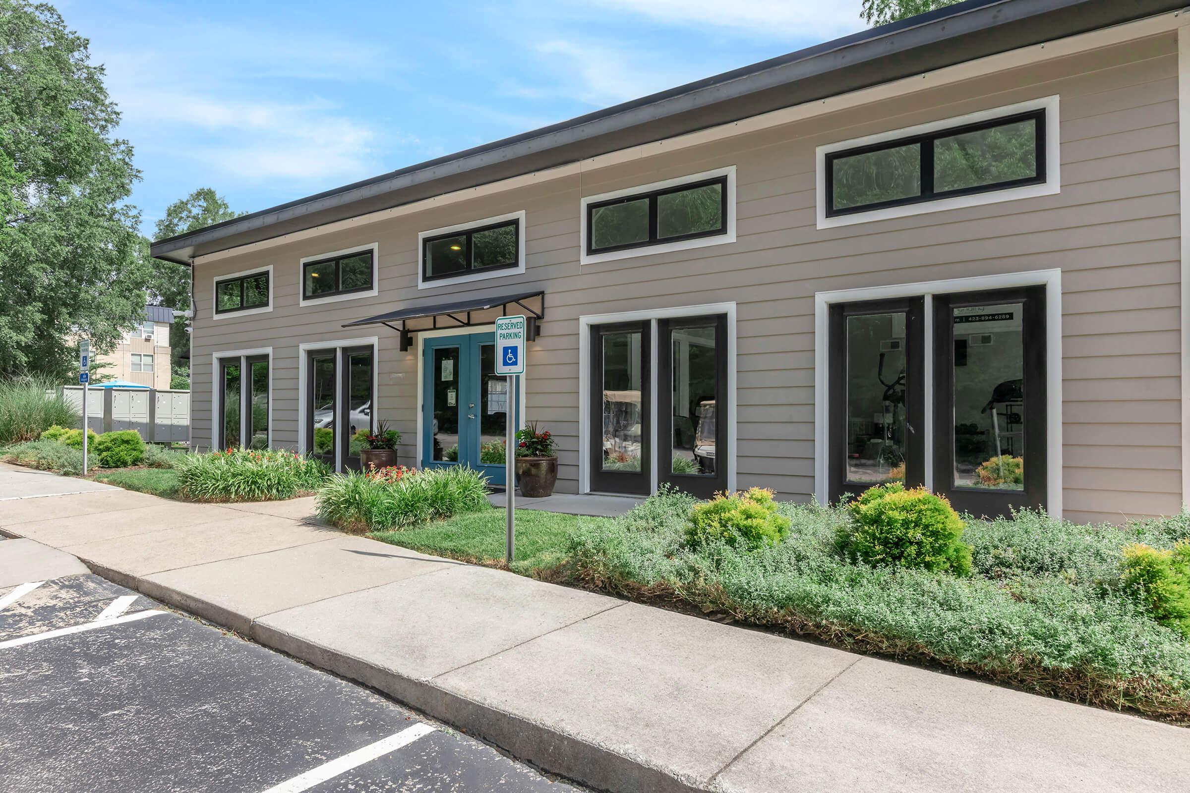A modern building with large windows and a blue door, surrounded by green landscaping. The structure features a light-colored exterior and is situated next to a parking area. The setting is bright and inviting, with trees in the background.