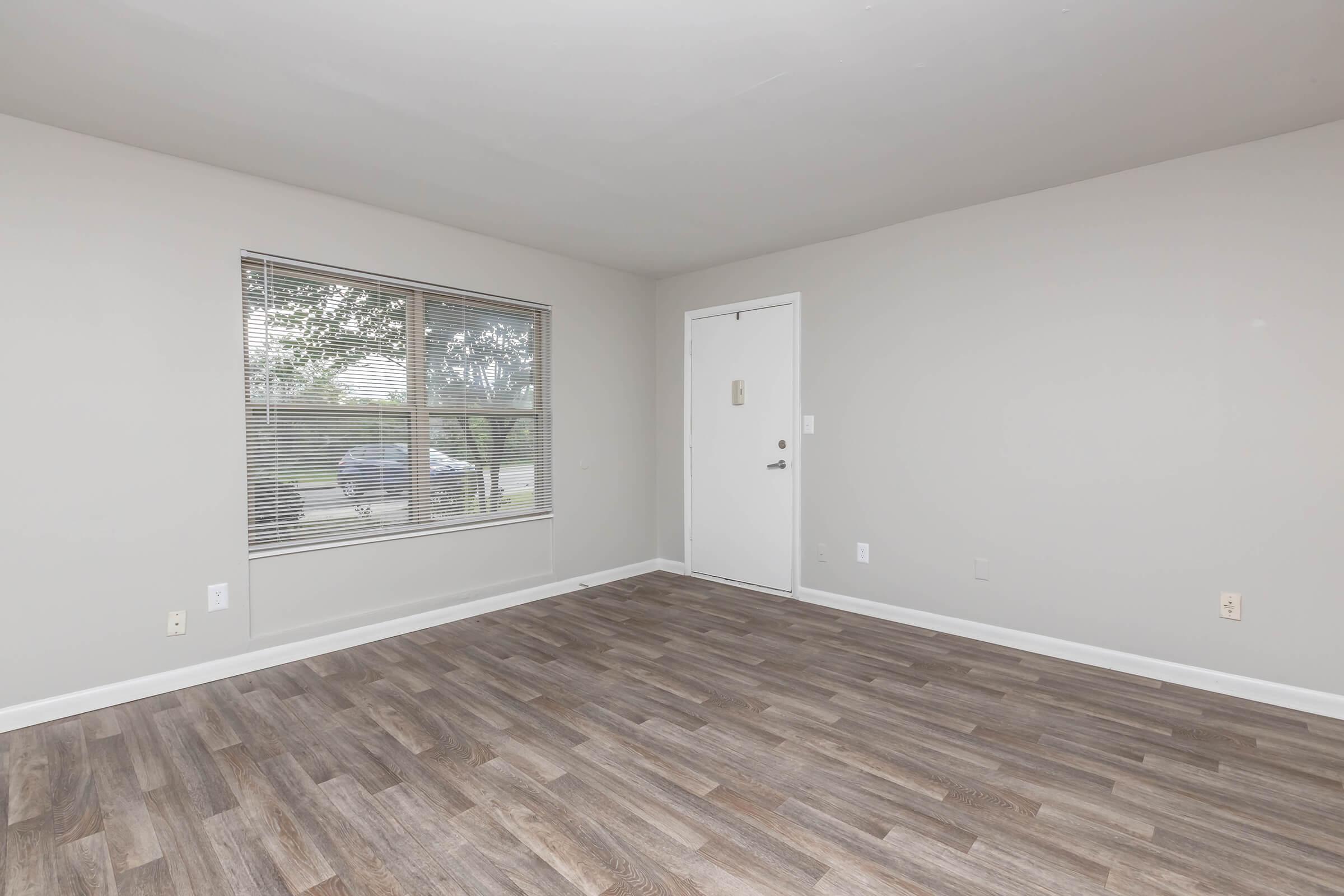 A spacious, empty room featuring light gray walls, a large window with blinds allowing natural light, and a door at the far side. The floor is covered with wooden laminate, creating a warm ambiance. The room lacks furniture, emphasizing its potential for customization.
