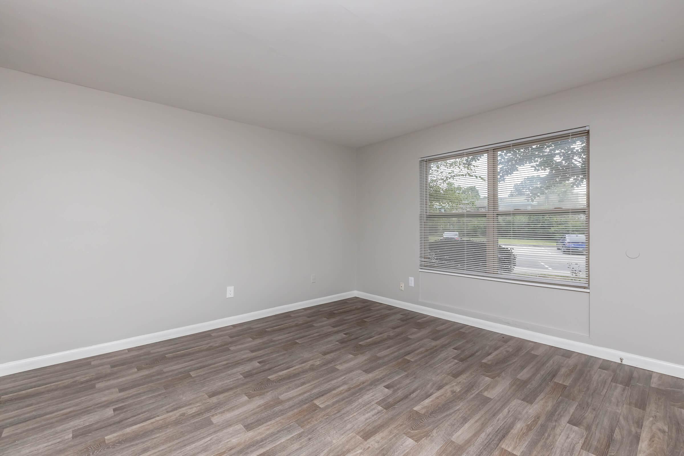 A spacious, empty room with light gray walls and a large window allowing natural light to enter. The floor features a wood-like laminate, and there are no furnishings present. Outside the window, a glimpse of greenery and a sidewalk are visible.