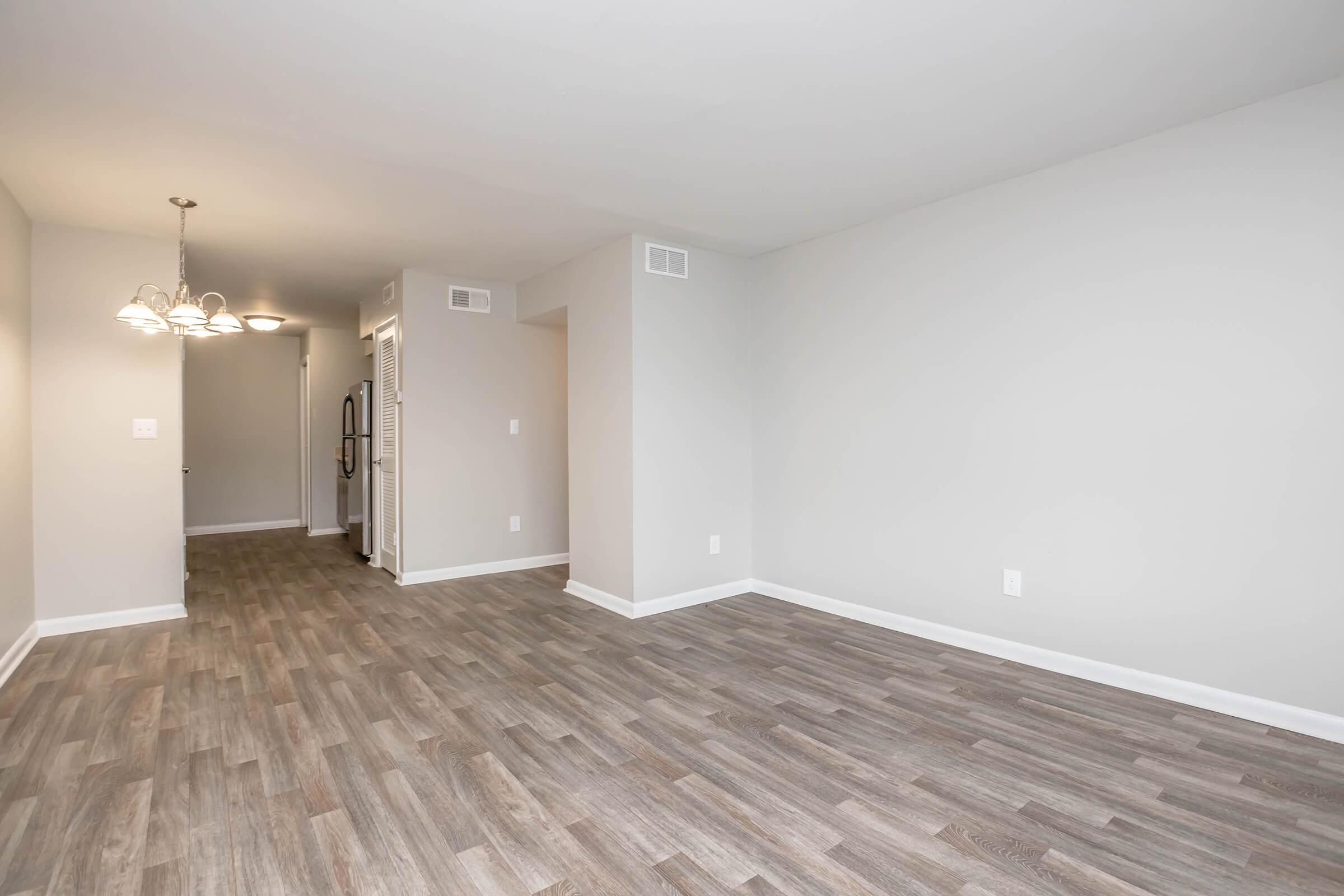 Empty, contemporary living space featuring light gray walls and wood-like flooring. A small dining area with a chandelier is visible, along with a doorway leading to another room. The kitchen is partially visible with appliances on display. The overall atmosphere is bright and spacious.