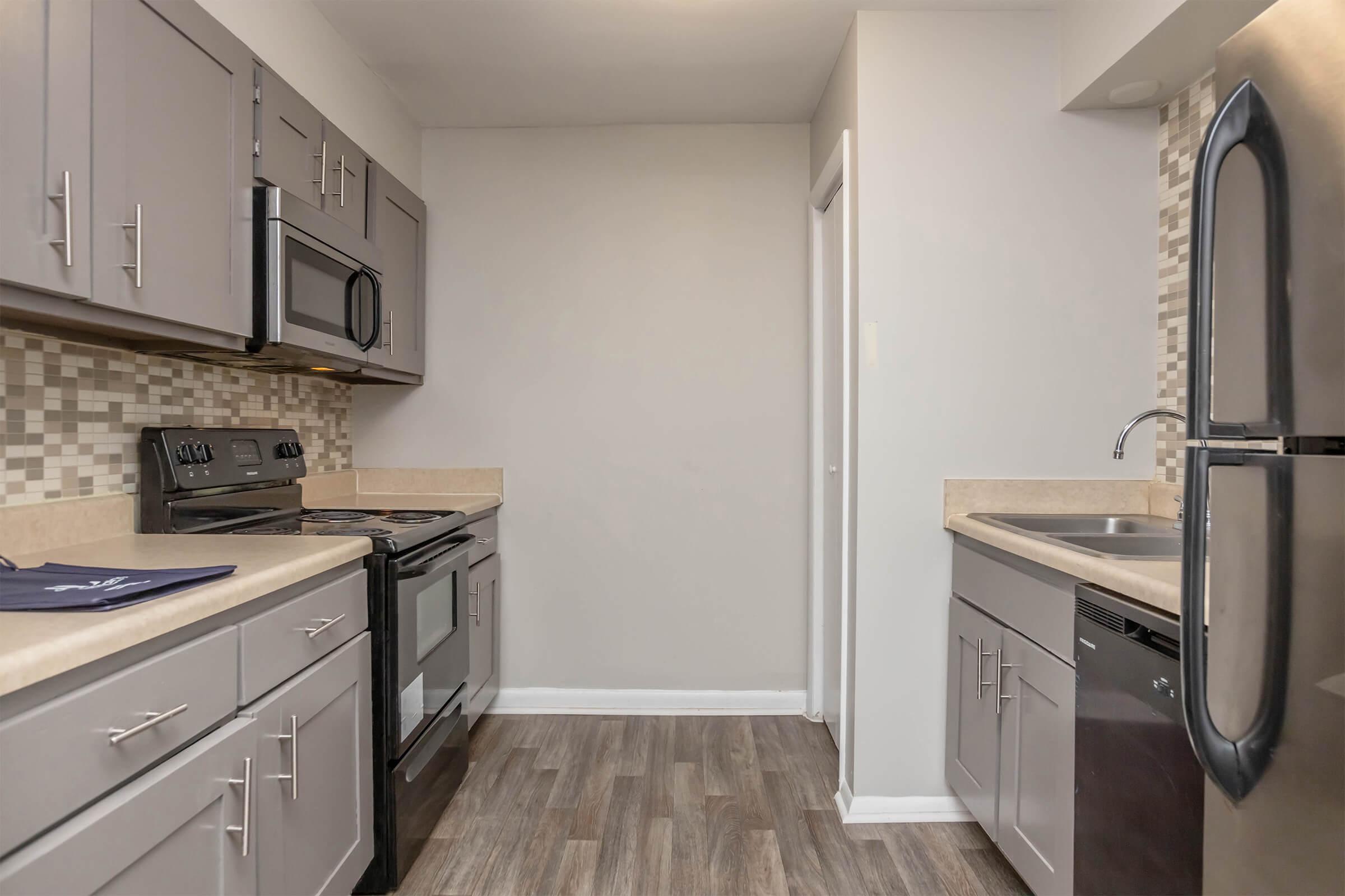Modern kitchen featuring sleek gray cabinets, stainless steel appliances including a refrigerator and oven, and a double sink. The countertops are light-colored, and there's a decorative tiled backsplash. The flooring is a wood-like laminate, creating a warm and inviting atmosphere.