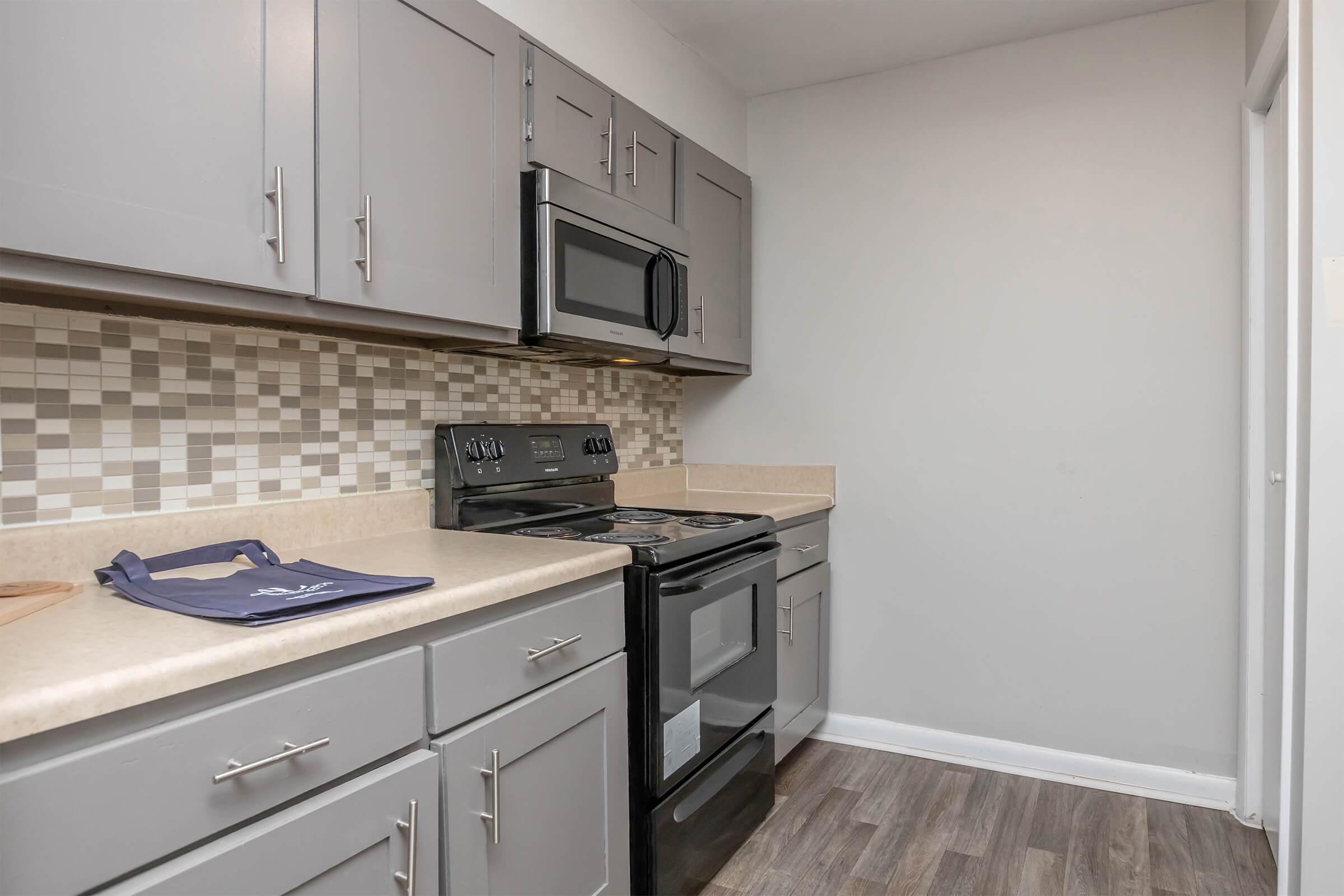 Modern kitchen featuring gray cabinets, a black stove and microwave, and a light countertop. The backsplash consists of a mosaic pattern in neutral tones. A dark blue kitchen towel is placed on the countertop, and the flooring is a dark laminate. The overall design is contemporary and inviting.