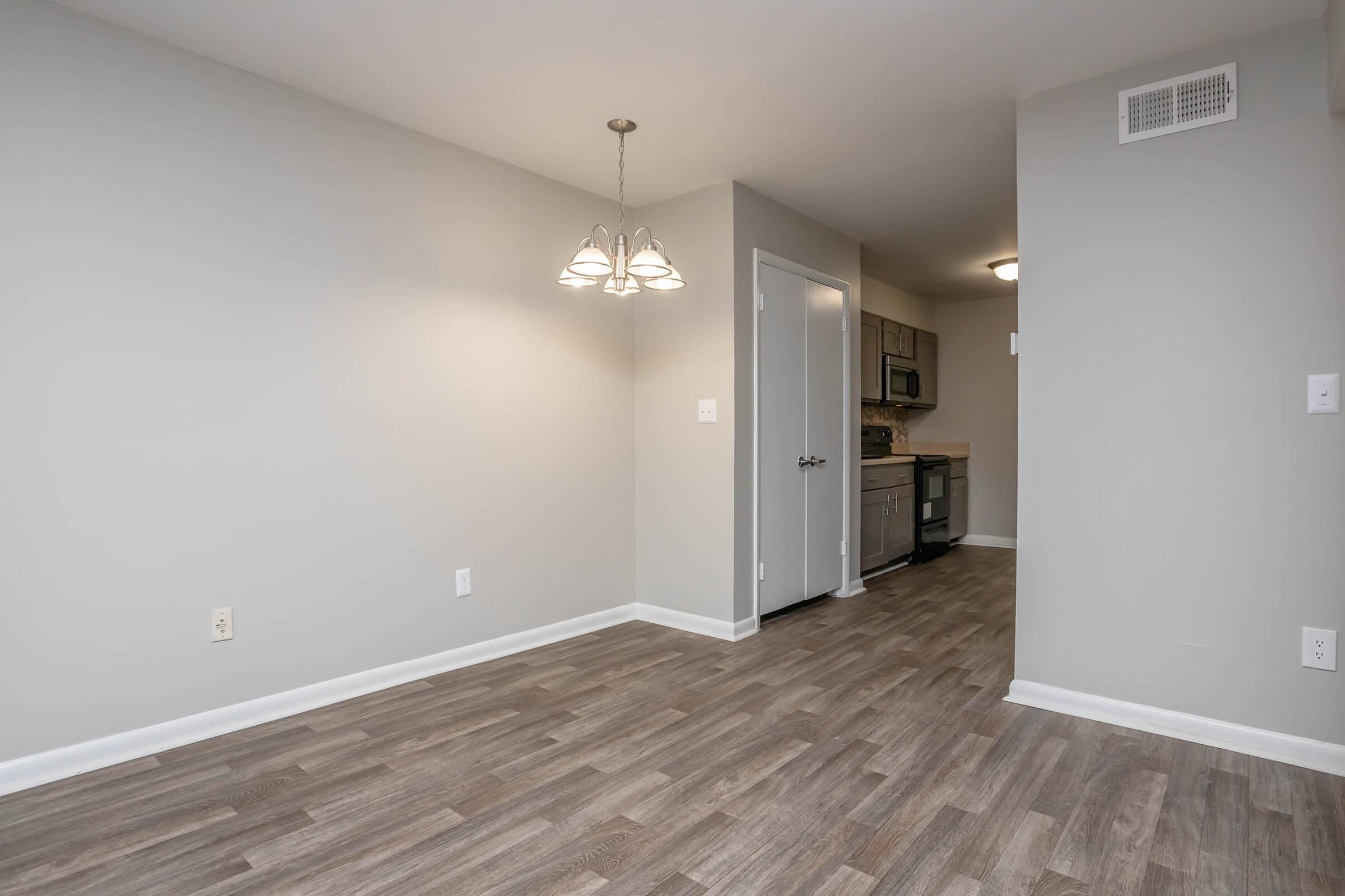 A modern, empty dining area with a light fixture hanging from the ceiling, featuring gray walls and light wood flooring. A doorway leads to a kitchen in the background, which is not fully visible. The space appears clean and freshly painted, inviting for furniture arrangement.