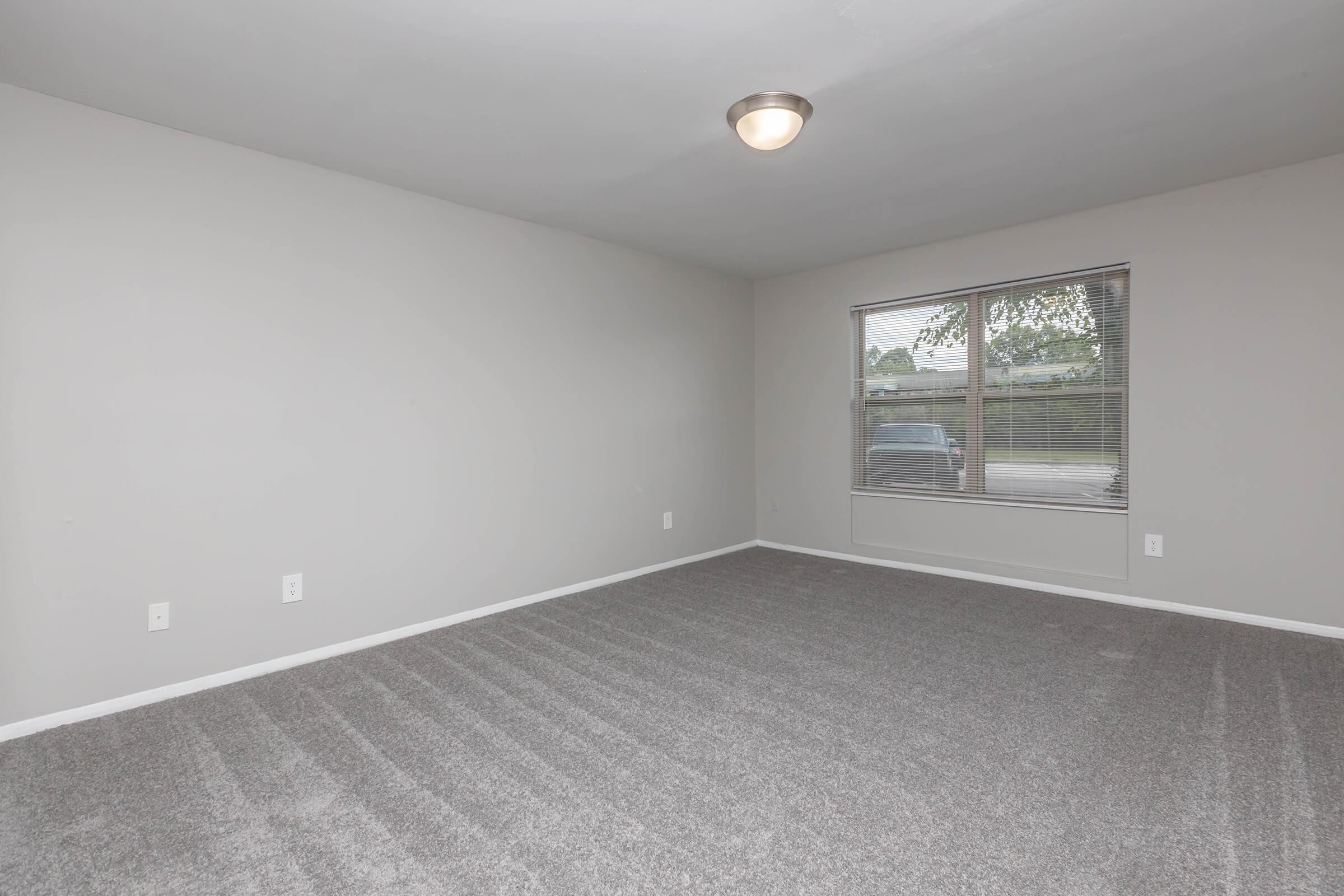 A spacious, empty room with light gray walls and new carpeting. A window allows natural light to enter, illuminating the unadorned space. The ceiling features a simple light fixture. The overall atmosphere is clean and ready for furnishing, ideal for a living area or office.