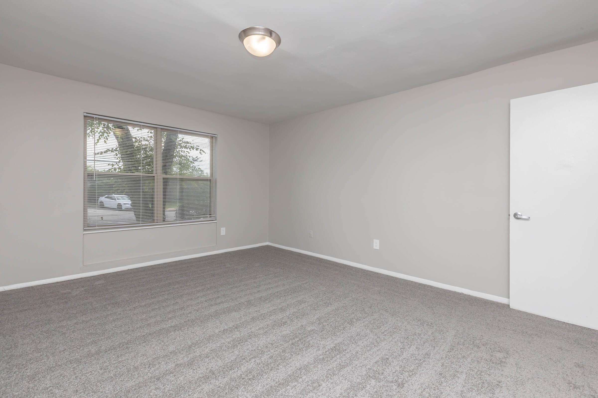 A spacious, empty room with light gray walls and a large window featuring blinds. The floor is covered in light gray carpet, and there is a ceiling light fixture. A door is visible on the right side of the image, and outside the window, a car can be seen parked.