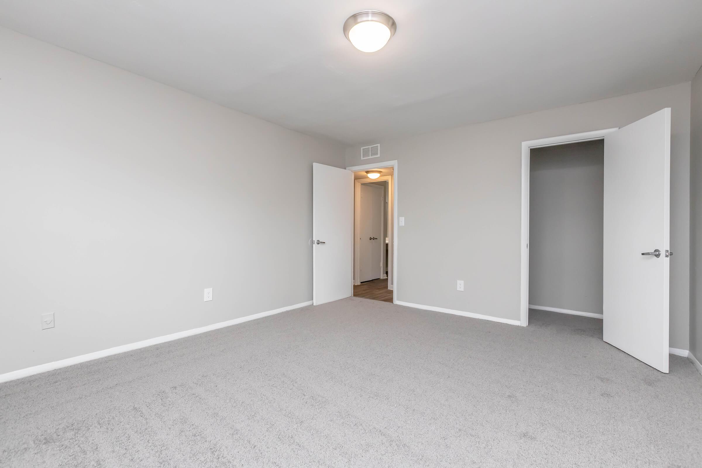 Empty room featuring light gray walls and gray carpet. Two white doors are visible, one slightly ajar, leading to another room or hallway. A ceiling light illuminates the space, which has no furniture or decorations, creating a clean and minimalist look.