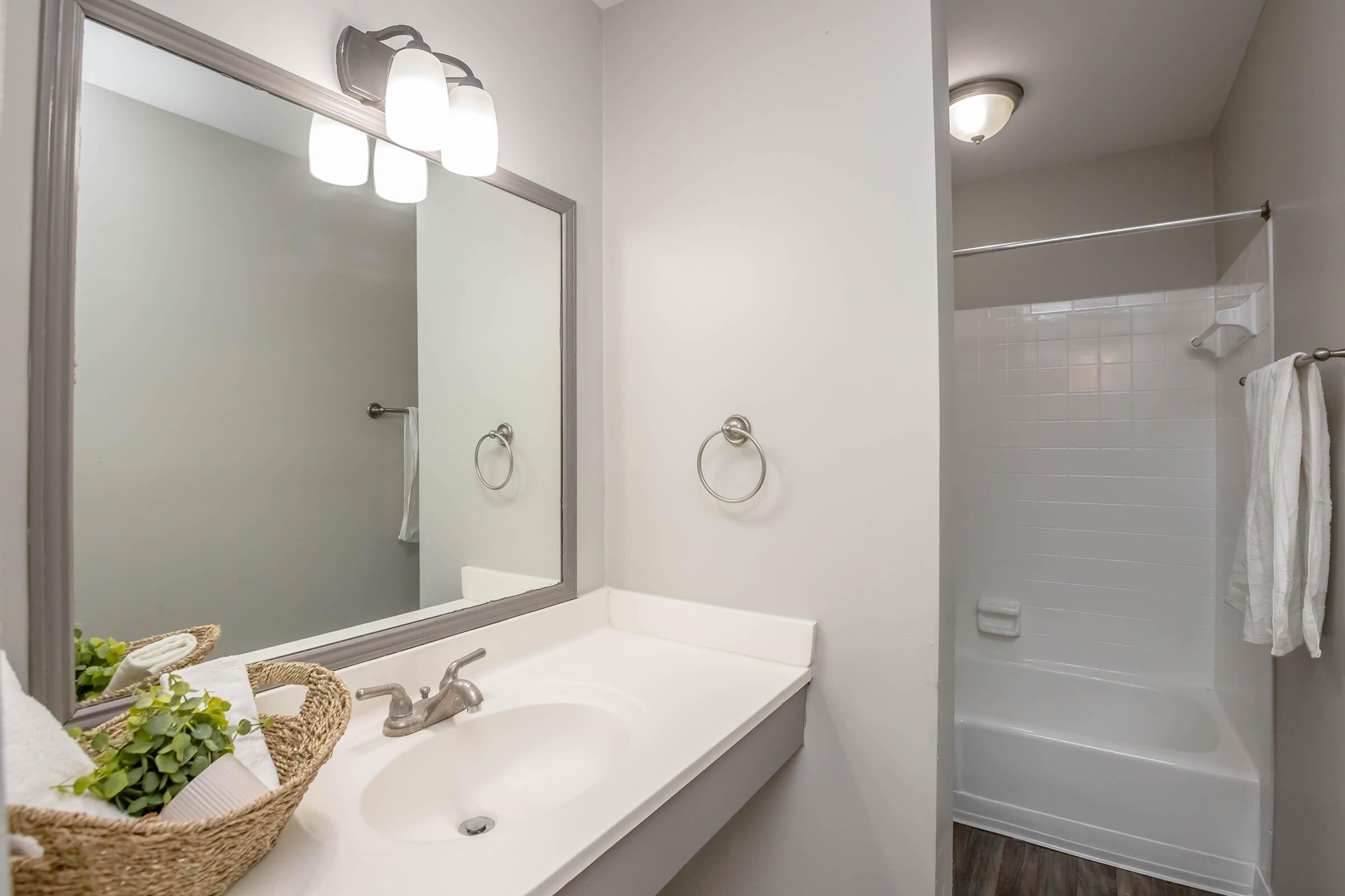 A modern bathroom featuring a white sink with silver fixtures, a large mirror framed in gray, and a shower area with a white tub. There’s a wall-mounted towel ring and white towels hanging. A decorative basket holds towels and greenery, enhancing the room's aesthetic.