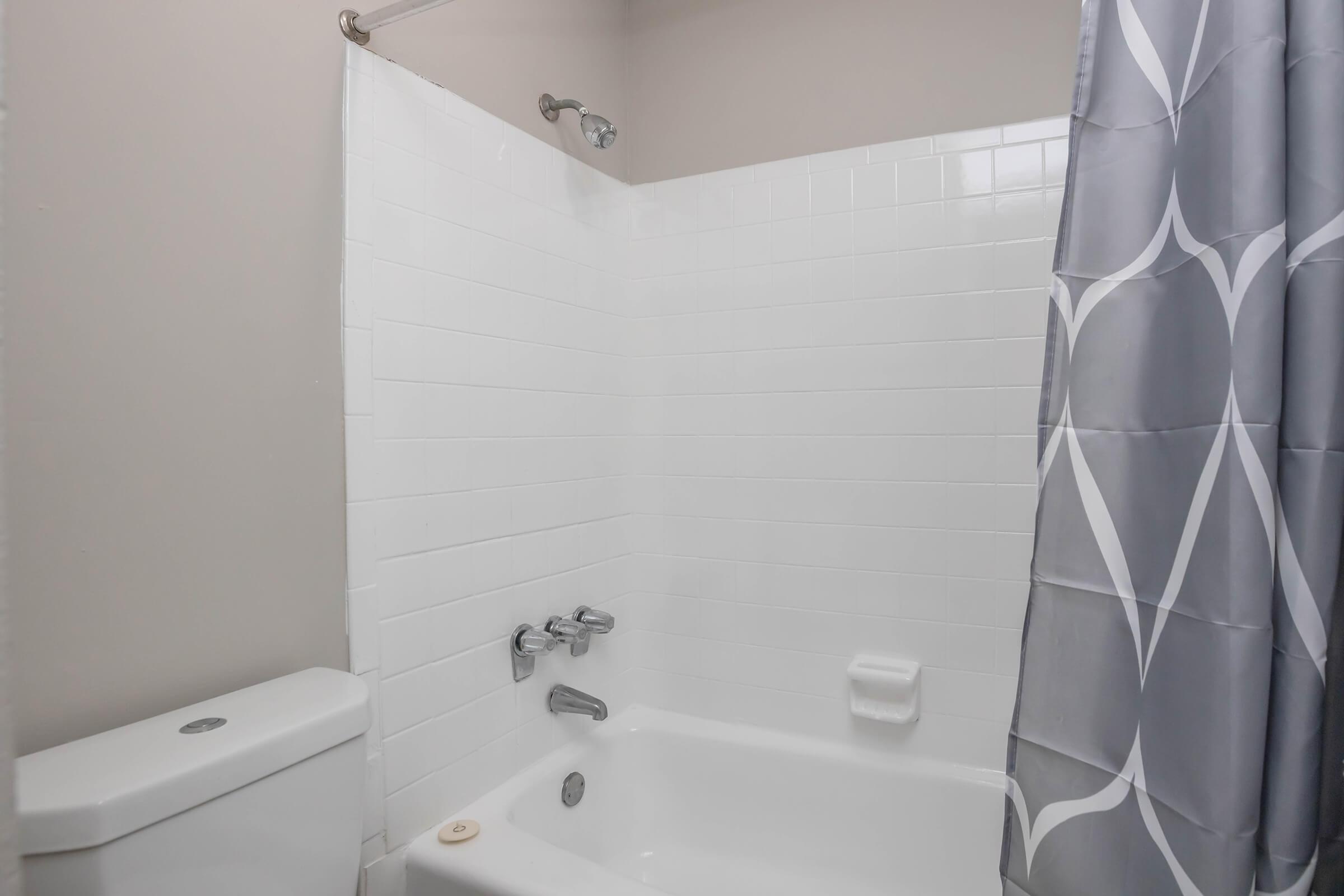 A clean bathroom featuring a white tiled shower area with a gray shower curtain, a bathtub, and a toilet. The shower fixture is visible on the wall, and the space is brightly lit, giving a fresh and modern appearance.