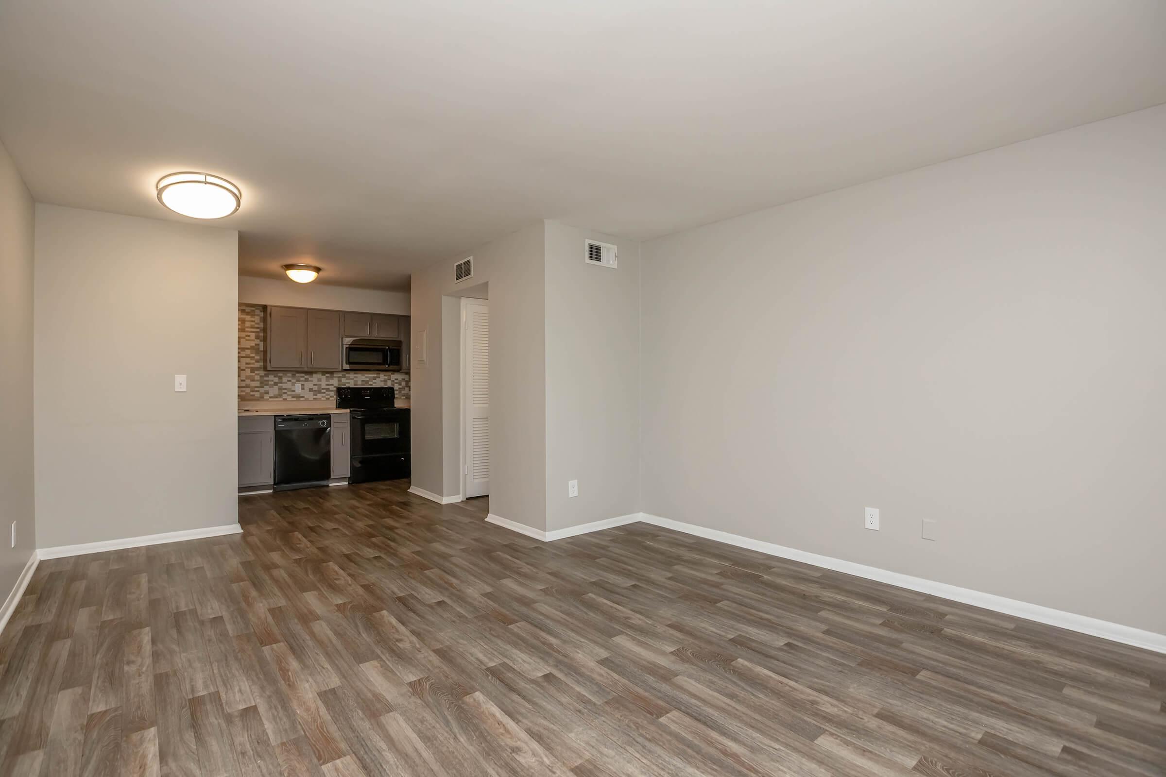 A spacious, empty living room featuring wood-style flooring, a light fixture on the ceiling, and an open layout leading to a modern kitchen. The kitchen has dark appliances and a tiled backsplash, with neutral gray walls enhancing the bright and airy atmosphere of the space.