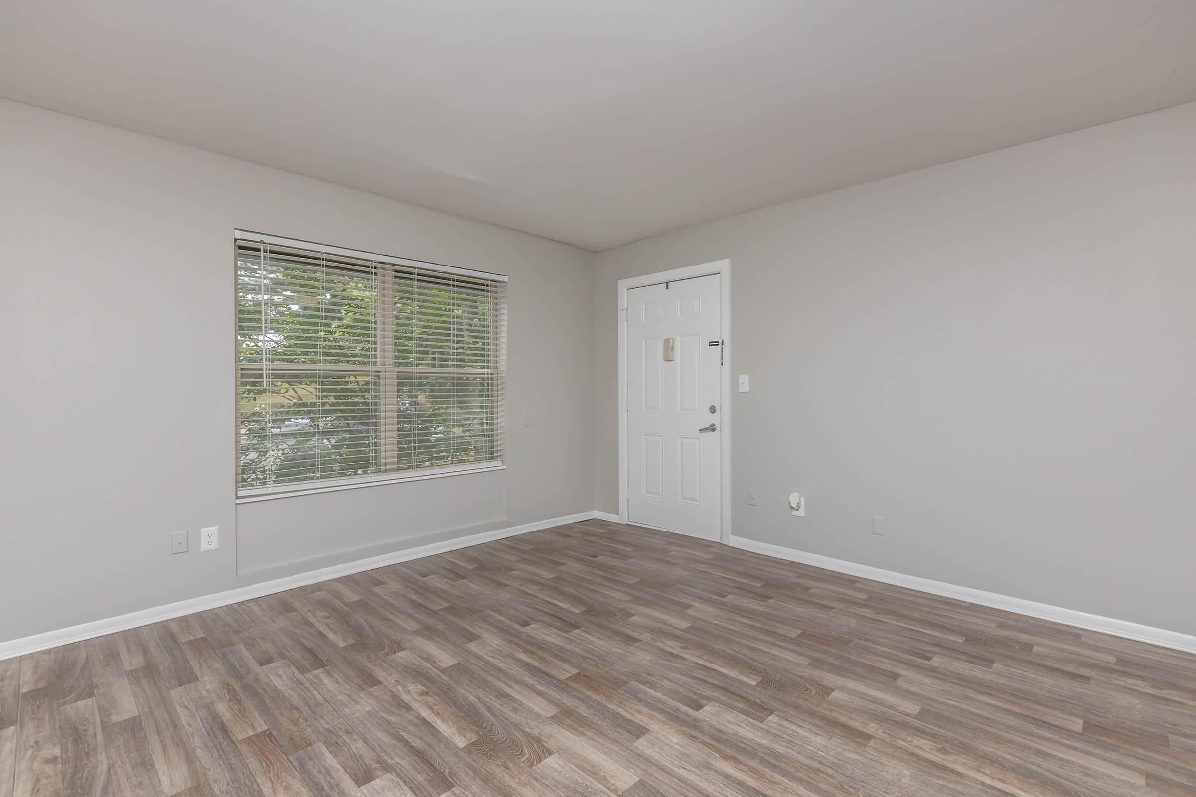 An empty room with light gray walls and a wooden floor. A window with blinds allows natural light, while a white door is visible on the right side. The space is unfurnished, creating an open and airy feel.