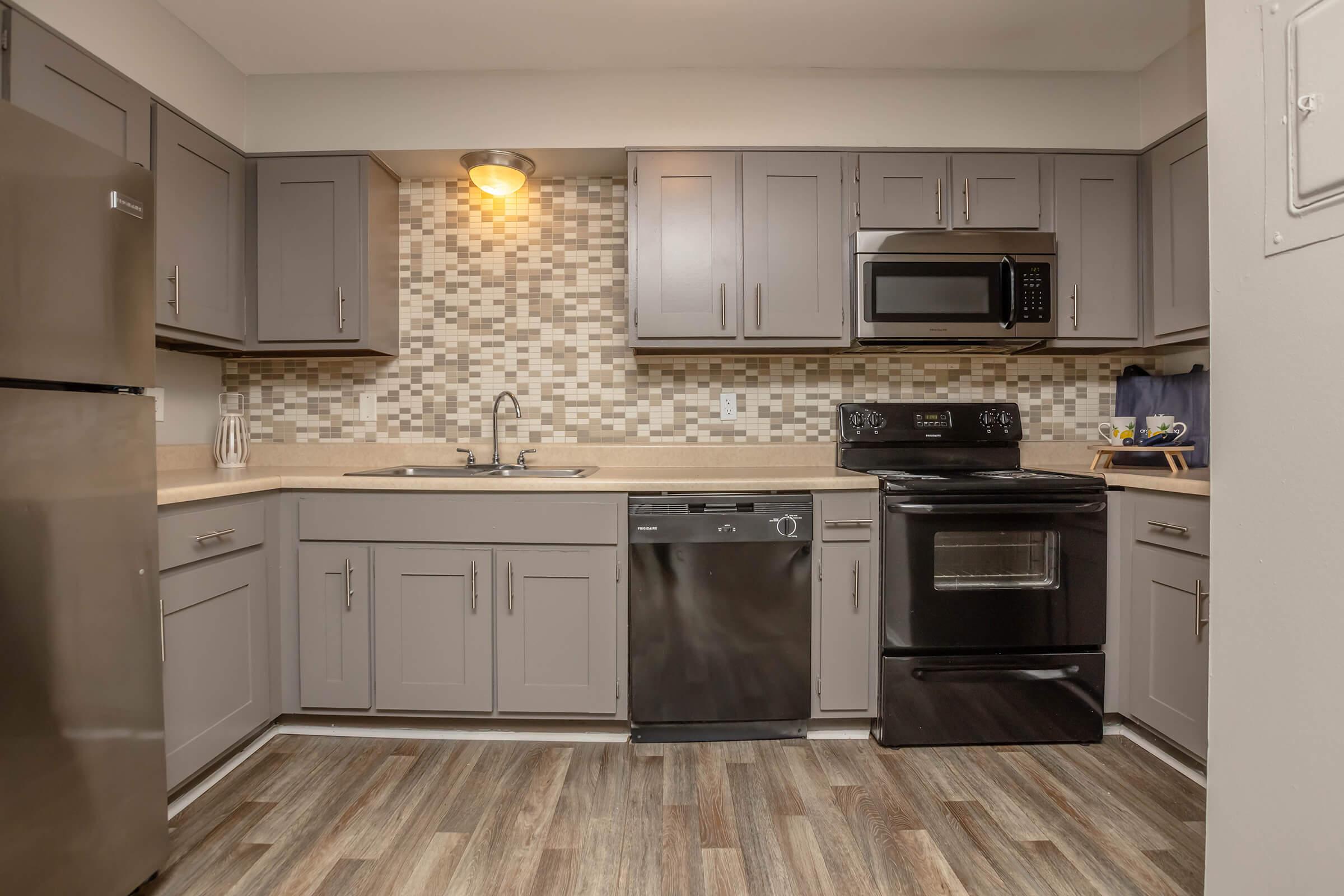 Modern kitchen featuring gray cabinets, stainless steel refrigerator, black dishwasher, and an oven with a microwave above. Countertops are light, complemented by a backsplash of mosaic tiles. Warm lighting adds a cozy ambiance, and the flooring is textured wood.