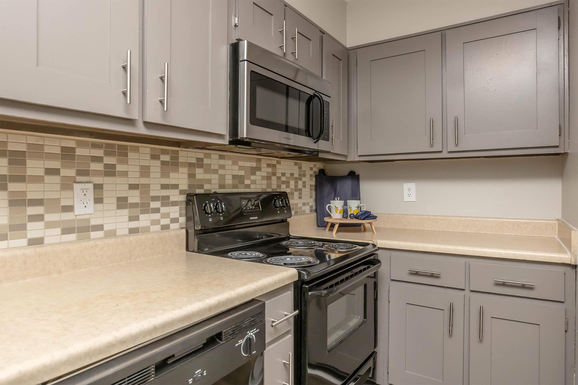 Modern kitchen featuring gray cabinets, a black stove, and a built-in microwave. The backsplash has a mosaic design in neutral tones. Countertops are light beige, and there are kitchen accessories on a stand. The overall design offers a contemporary and functional cooking space.