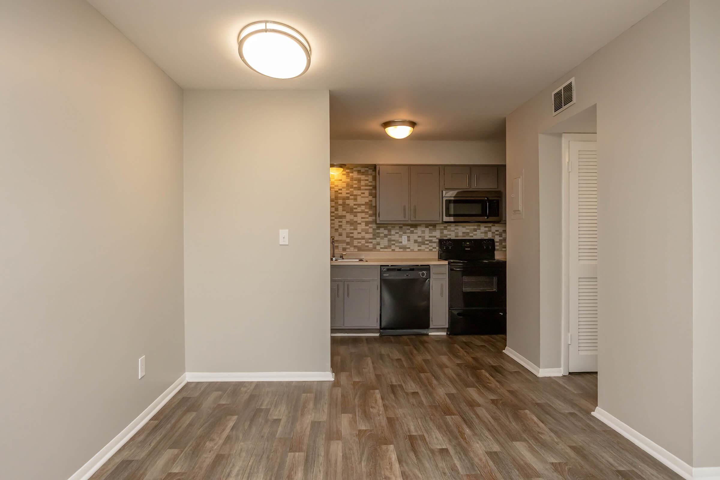 A view of a modern kitchen and living space featuring light gray walls, wooden flooring, and a ceiling light fixture. The kitchen includes gray cabinets, a black stove, a dishwasher, and a tiled backsplash. A doorway is visible leading to another area in the apartment.