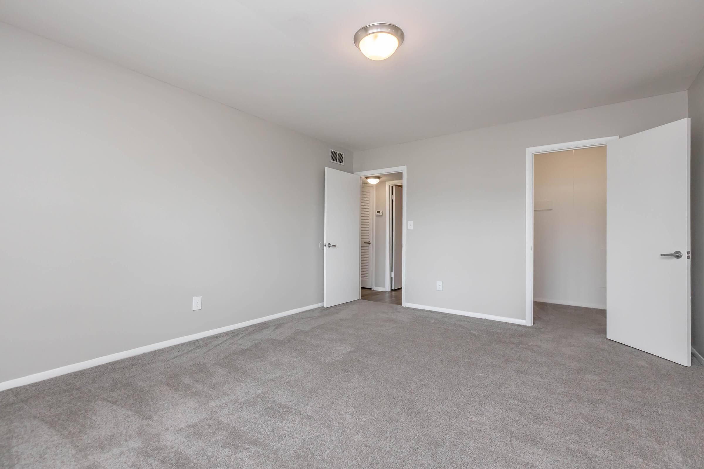 Spacious, empty room with light gray walls and soft, carpeted flooring. The room features a ceiling light fixture and two doors leading to other spaces, creating an inviting atmosphere. Ideal for personalizing with furniture and decor.