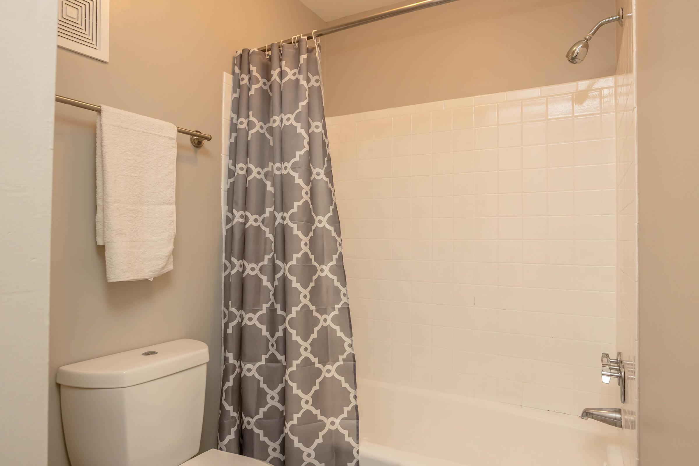 A bathroom featuring a white bathtub with a shower. There is a gray and white patterned shower curtain, a white towel hanging on a towel rack, and a toilet beside the bathtub. The walls are painted in a neutral tone.
