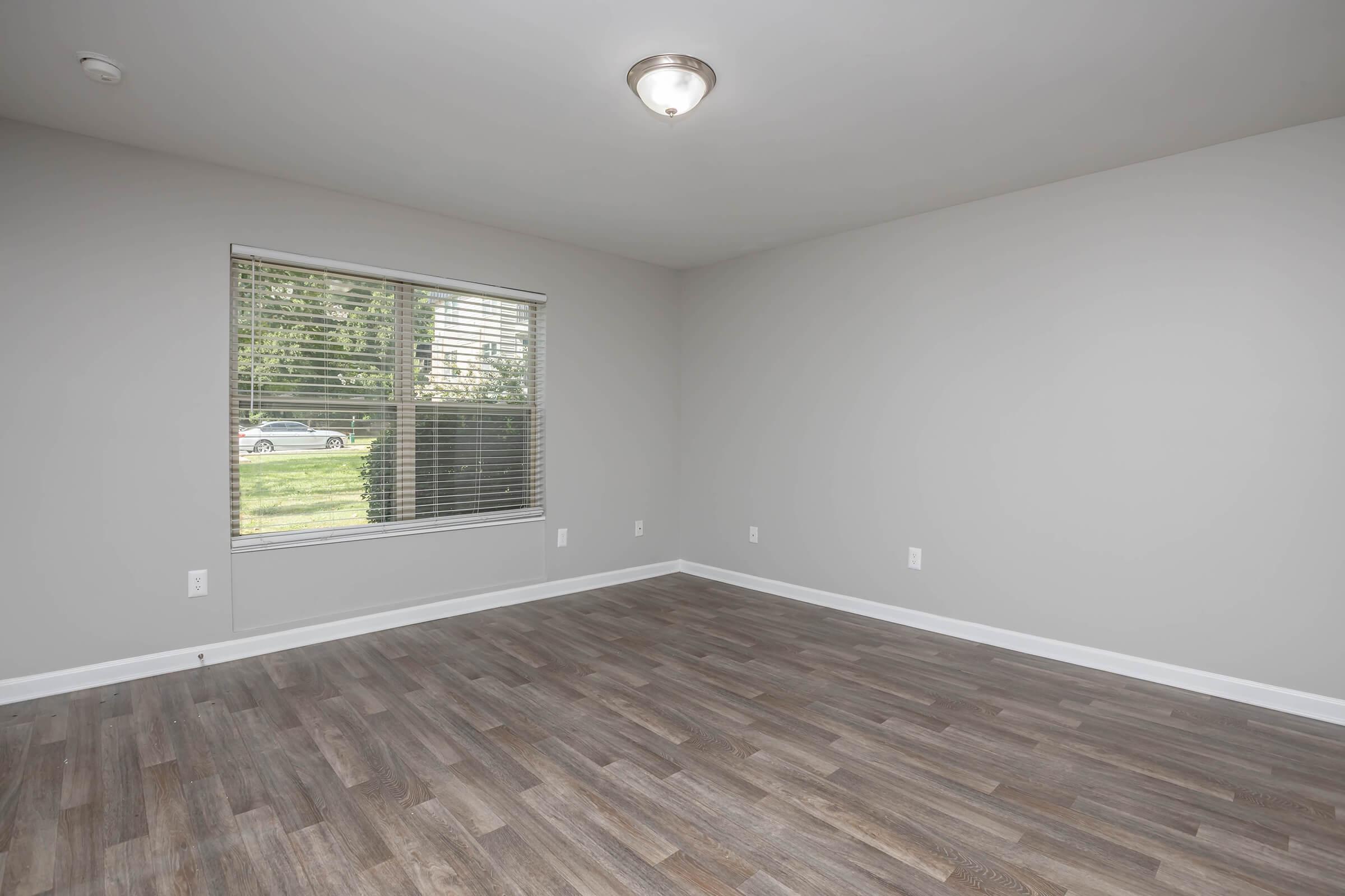 A vacant room with light gray walls and a wooden floor. There is a window with blinds on one wall, allowing natural light to enter, and a ceiling light fixture. The space appears clean and ready for furnishings, with no decorations or furniture present.