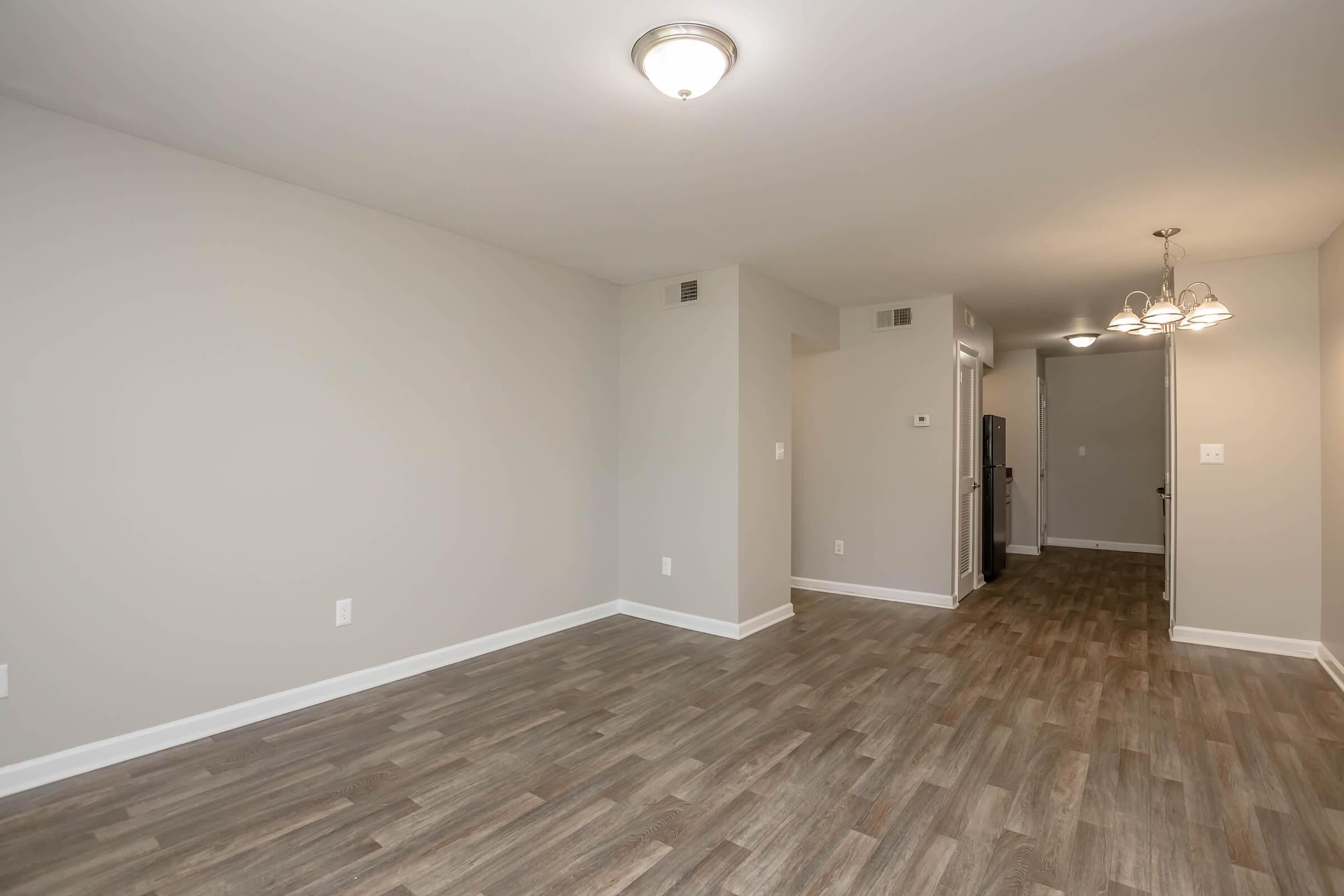 A spacious and well-lit living area with neutral gray walls and laminate flooring. There are doorways leading to other rooms, and a light fixture on the ceiling. A glimpse of a kitchen can be seen through an open doorway, with additional lighting from a chandelier in the background.