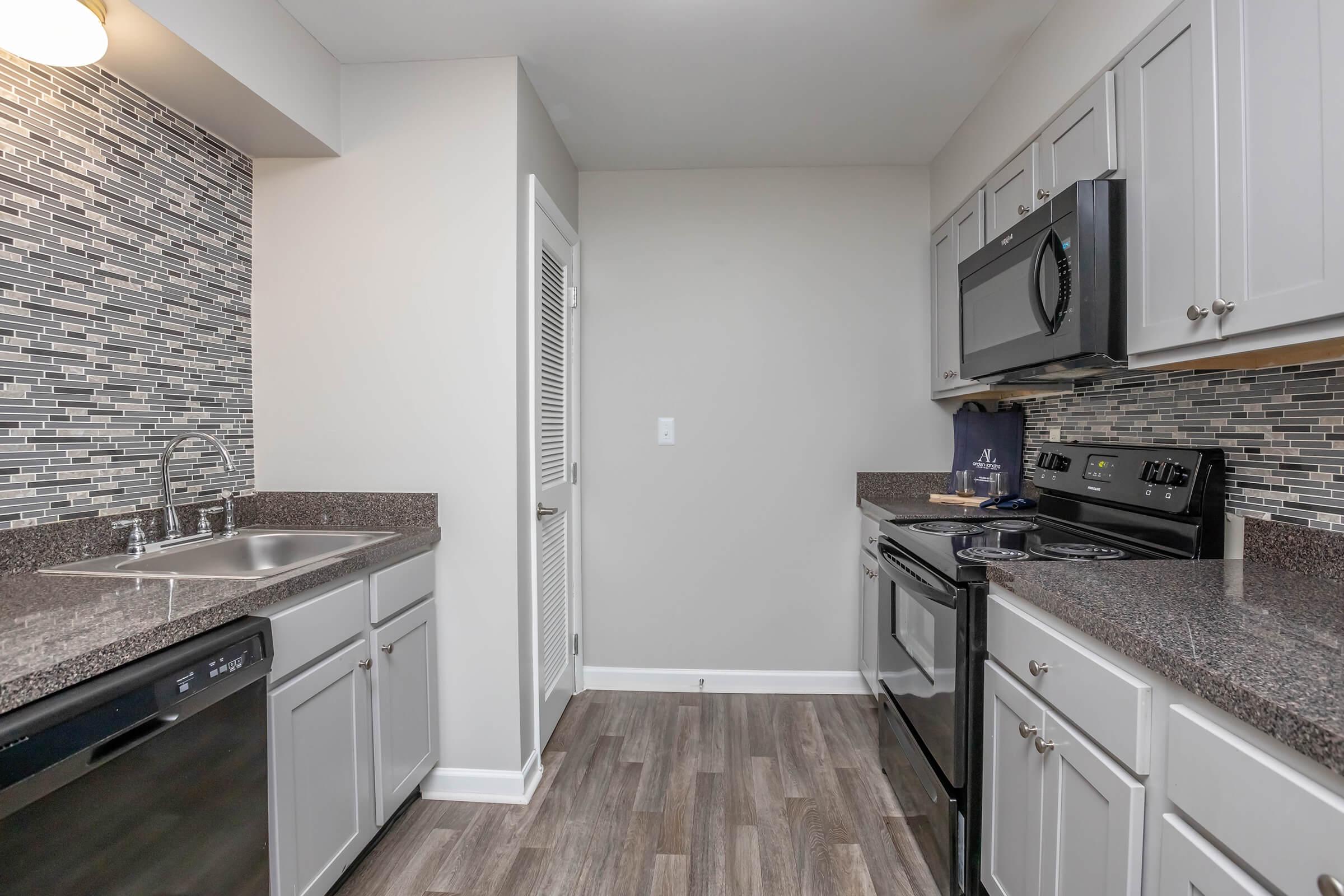 Modern kitchen featuring gray cabinets, granite countertops, and stainless steel appliances. The space includes a stainless steel sink, dishwasher, oven, and microwave, with a decorative tile backsplash. The flooring is wood-like, and the walls are painted in a neutral color, creating a bright and inviting atmosphere.