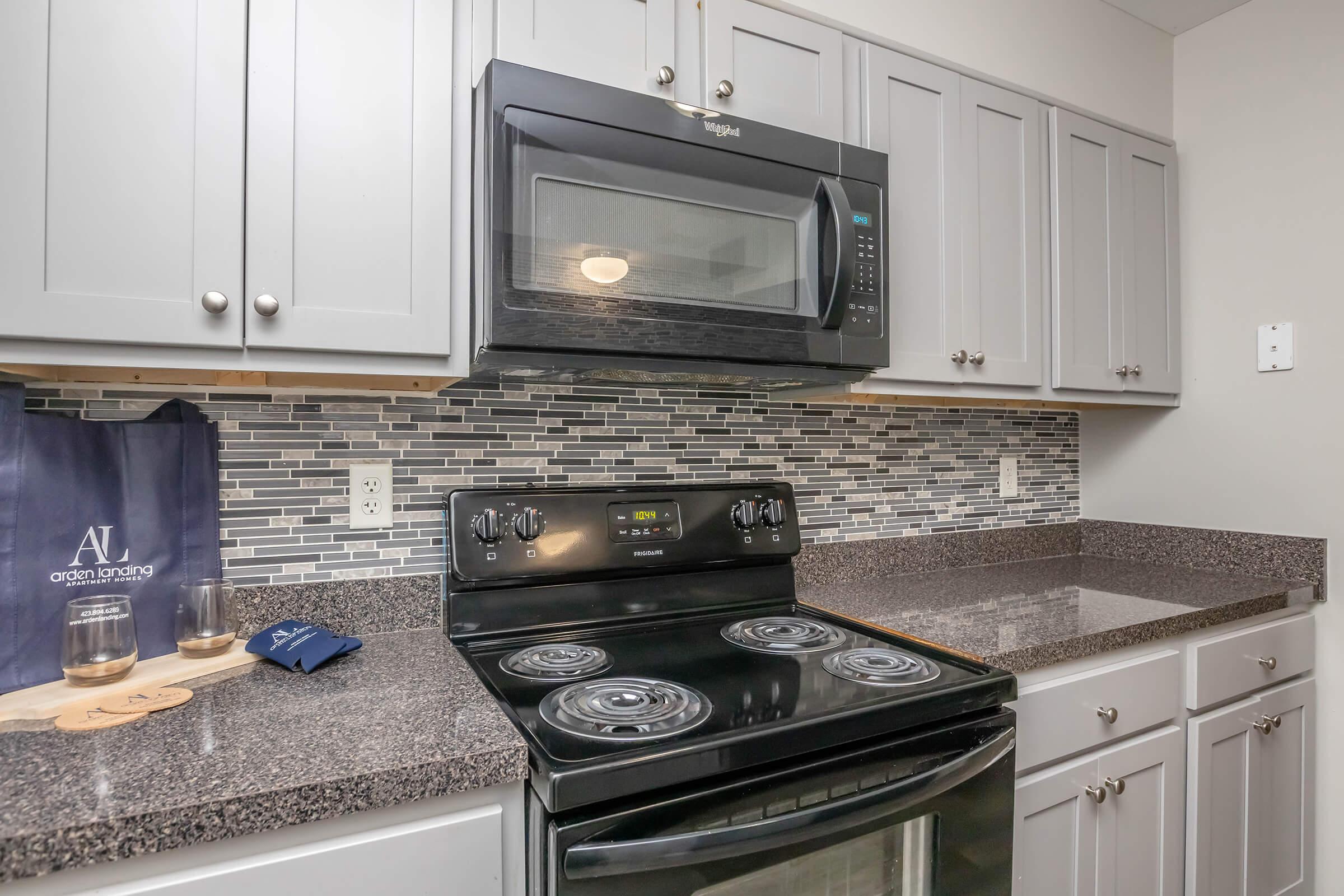A modern kitchen featuring dark granite countertops, a black stove, and a built-in microwave. The cabinets above are light gray with a mosaic tile backsplash. A shopping bag is on the counter, along with glasses and a coaster, creating a warm and inviting atmosphere.