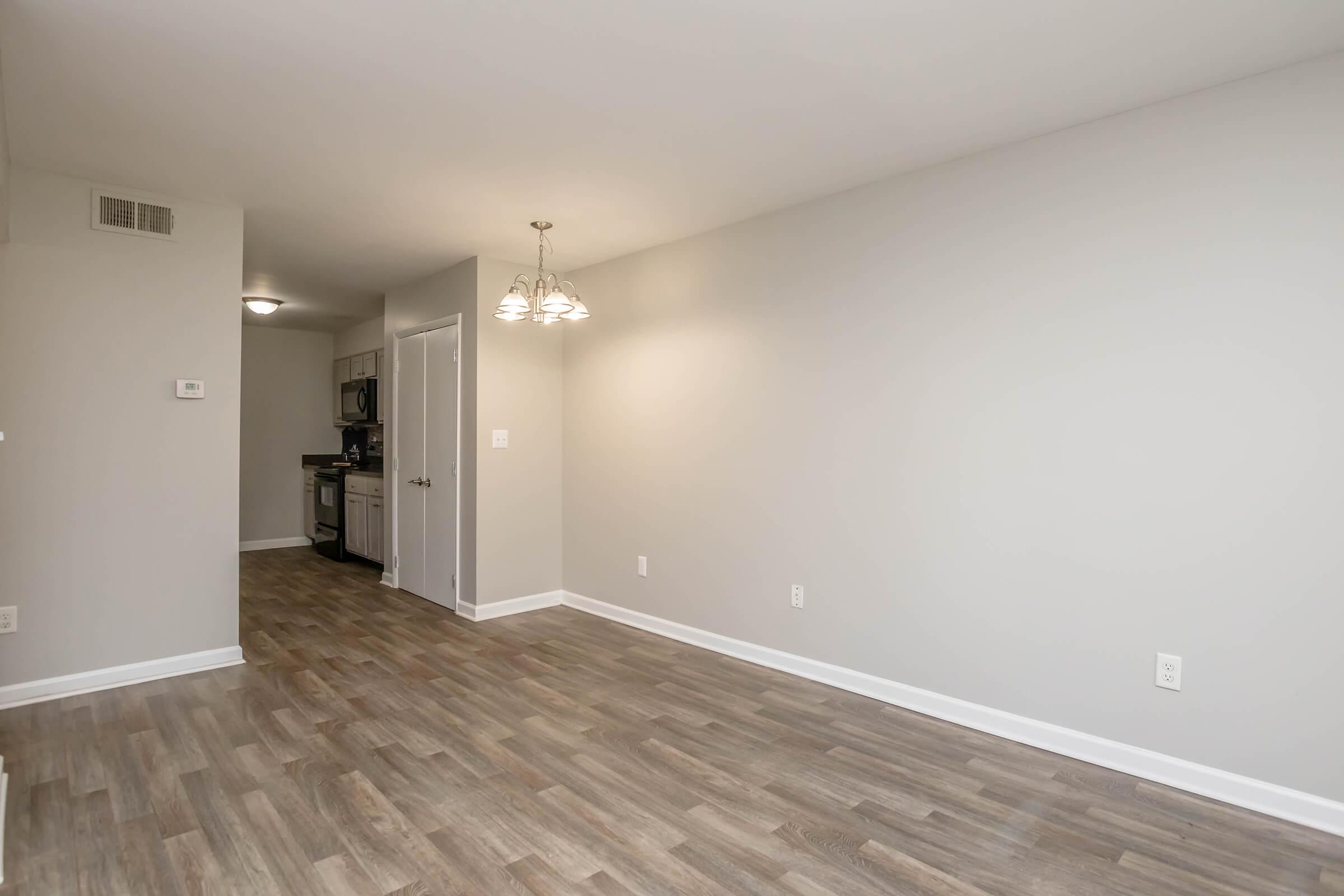 A spacious, well-lit living area with light gray walls and a wood-look floor. In the background, there’s a doorway leading to a kitchen with modern appliances. A small, pendant light hangs from the ceiling, providing illumination. The room is empty, offering a blank canvas for potential furniture placement.