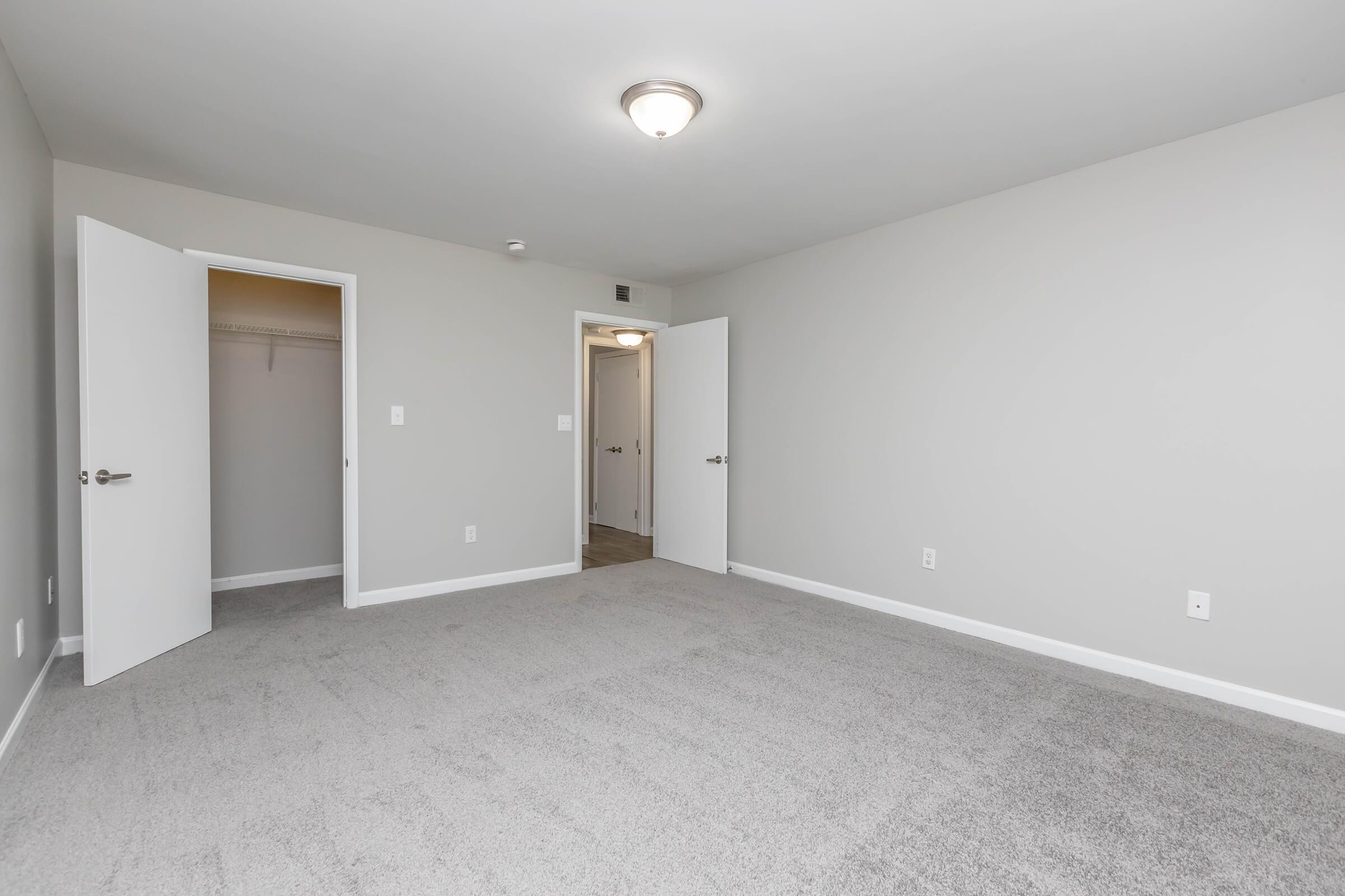 A spacious, empty room with light gray walls and carpeted flooring. Two white doors are open, revealing a closet and another doorway. The room is well-lit by a ceiling light fixture, creating a bright and inviting atmosphere. The overall design is modern and minimalistic.