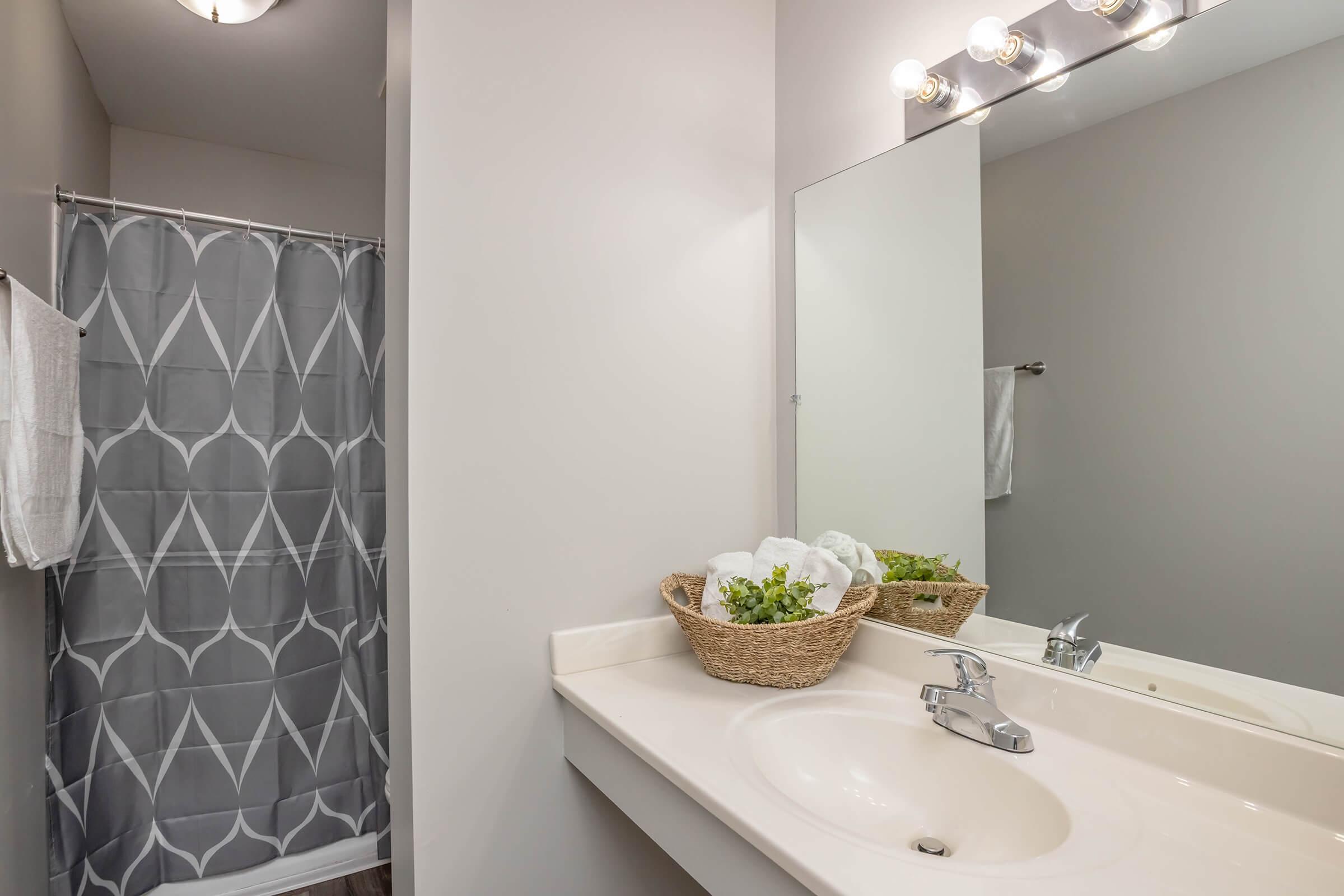 A bright bathroom featuring a light gray shower curtain with a geometric pattern, a large mirror above the sink with four light fixtures, a white countertop, and a decorative basket with white towels and greenery on the counter. There is a white towel hanging on the wall.