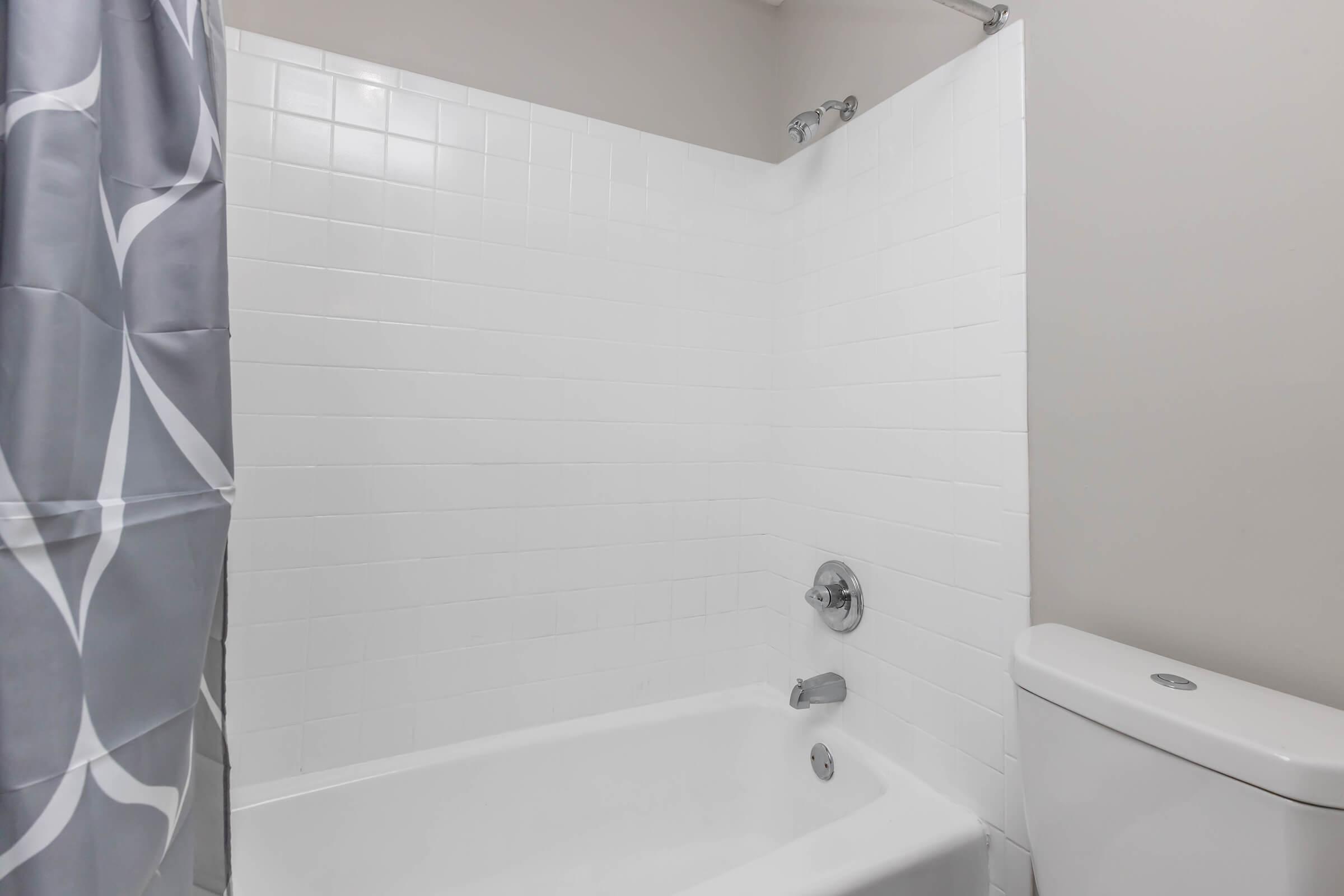 A clean bathroom featuring a white tiled shower and bathtub, with a silver showerhead and faucet. The bathtub is white and the adjacent toilet is also white. A gray shower curtain with a subtle pattern hangs nearby, adding a touch of decor to the minimalist space. The walls are painted in a neutral tone.