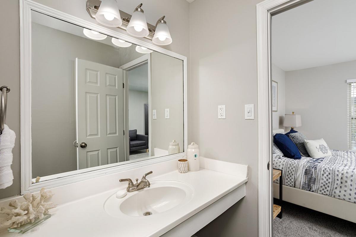 A bright, modern bathroom featuring a white countertop sink, a large mirror, and stylish lighting. The bathroom opens to a softly decorated bedroom with a bed adorned with blue and gray bedding. A towel rack and toiletries are neatly arranged, creating a clean and inviting atmosphere.