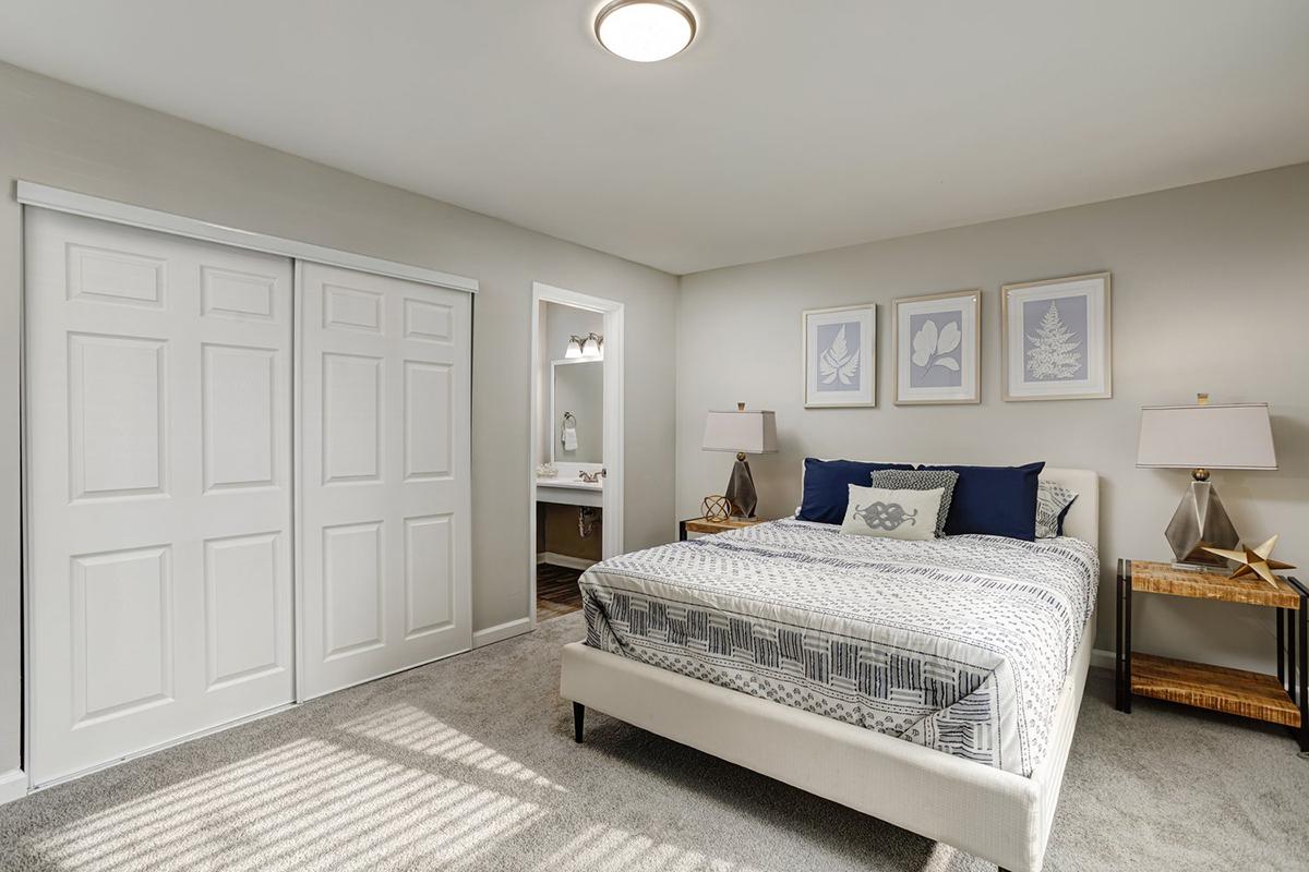 A neatly arranged bedroom featuring a light gray bed with decorative pillows, two bedside lamps, and framed botanical prints on the wall. There is a closet with sliding doors, and a door leading to an adjoining bathroom visible in the background. The room has neutral-colored walls and carpeting, creating a calm atmosphere.