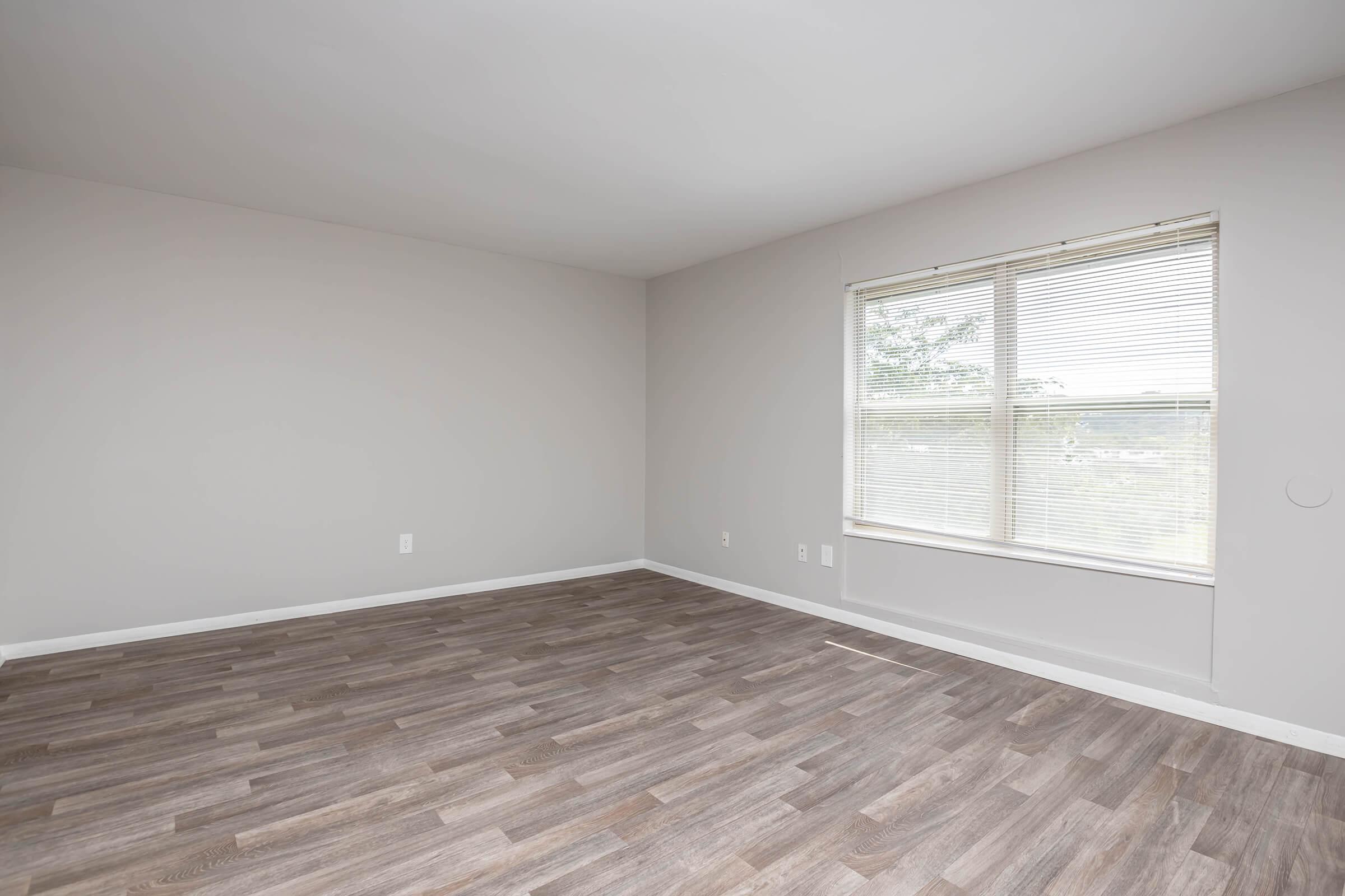 Empty room with light gray walls and large window allowing natural light. The floor is covered with brown wooden laminate. There are no furniture or decorations, creating a spacious and minimalist atmosphere.