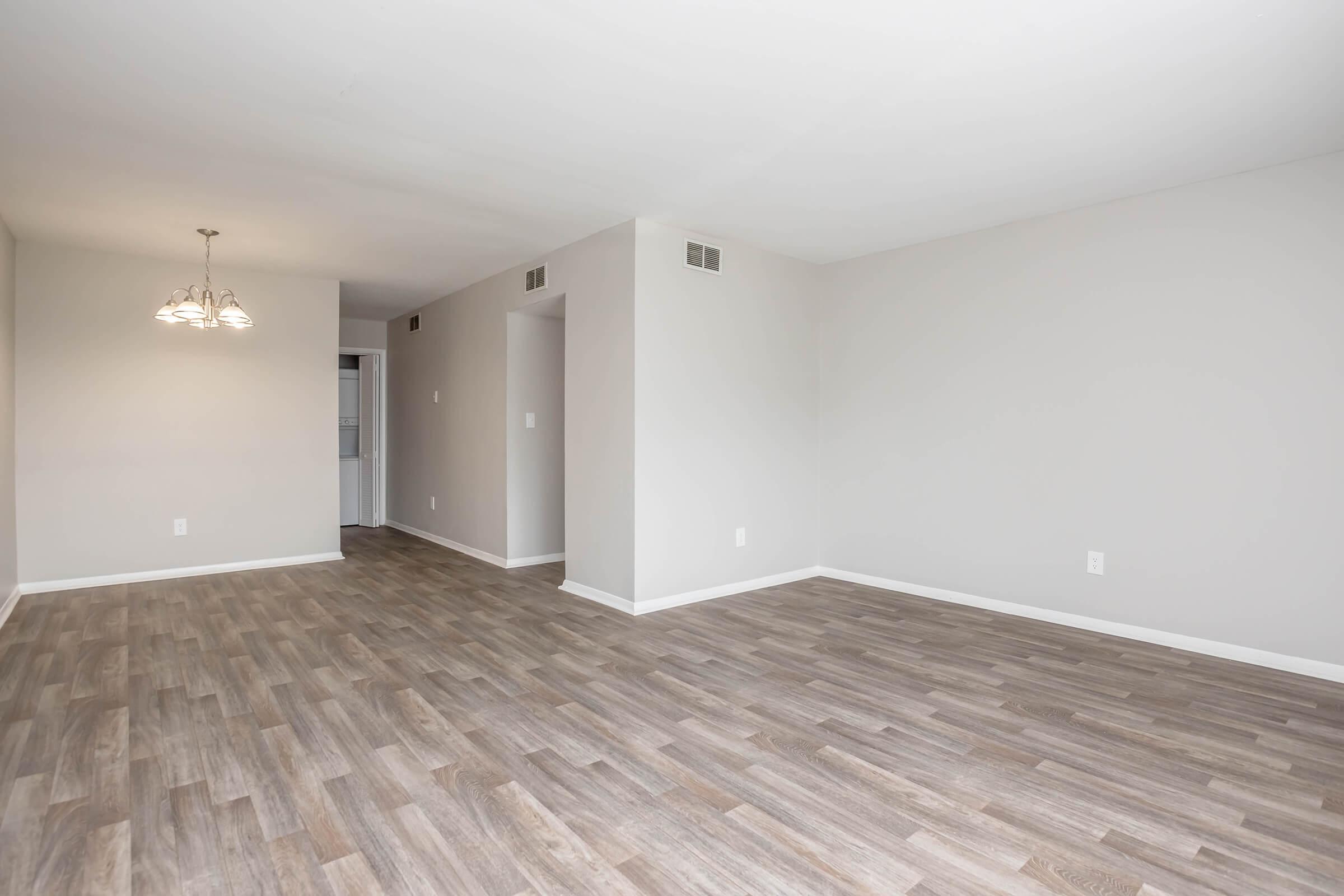 A spacious, empty room with light-colored walls and a wooden floor. A small chandelier hangs from the ceiling, and there is a doorway leading to another room. Natural light fills the area, creating a bright, open atmosphere.