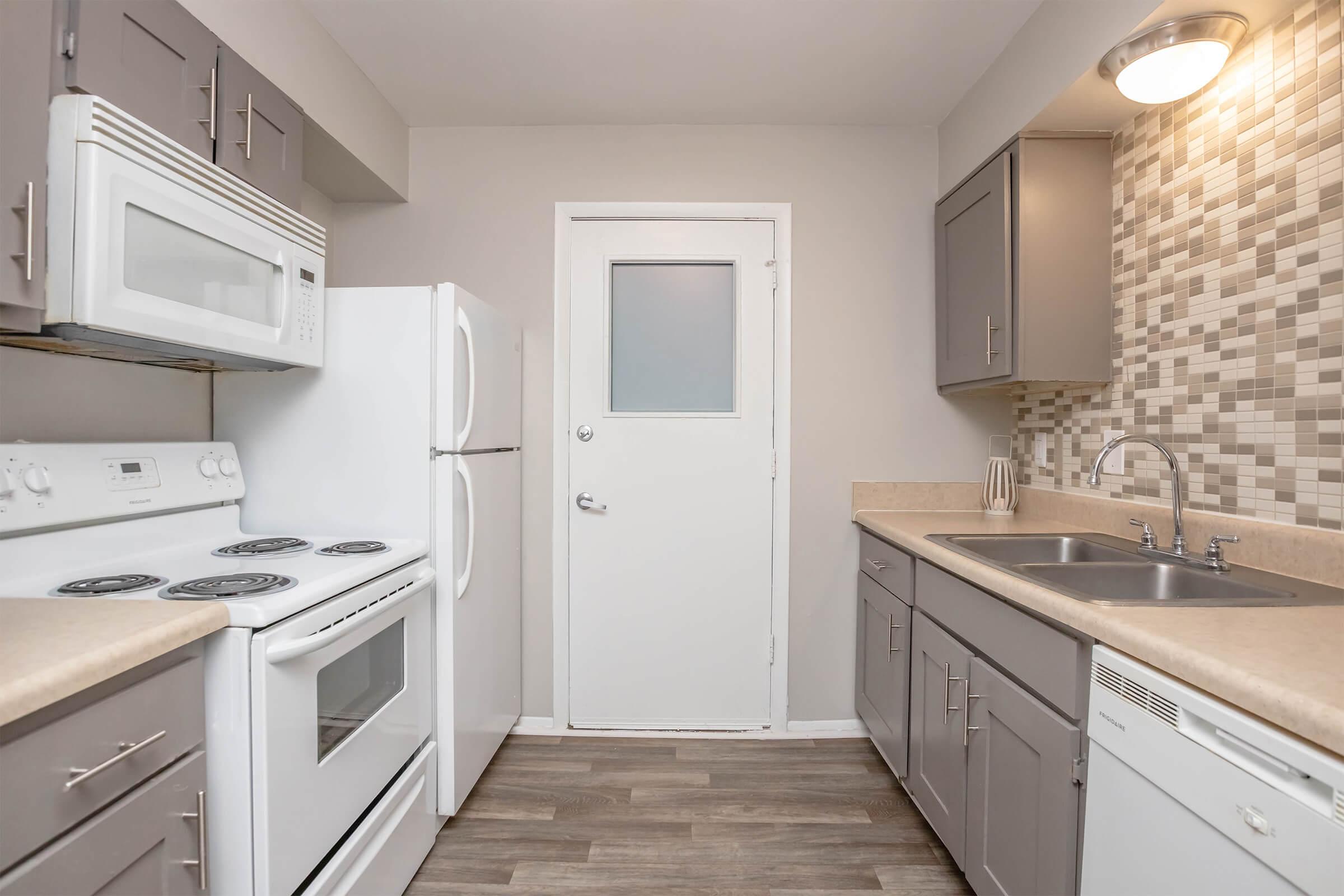 Modern kitchen featuring white appliances, including a stove, microwave, and refrigerator. The counters are light-colored with gray cabinets beneath. The backsplash consists of stylish mosaic tiles in neutral tones. A door leads to another area, and the floor is covered with laminate planks.